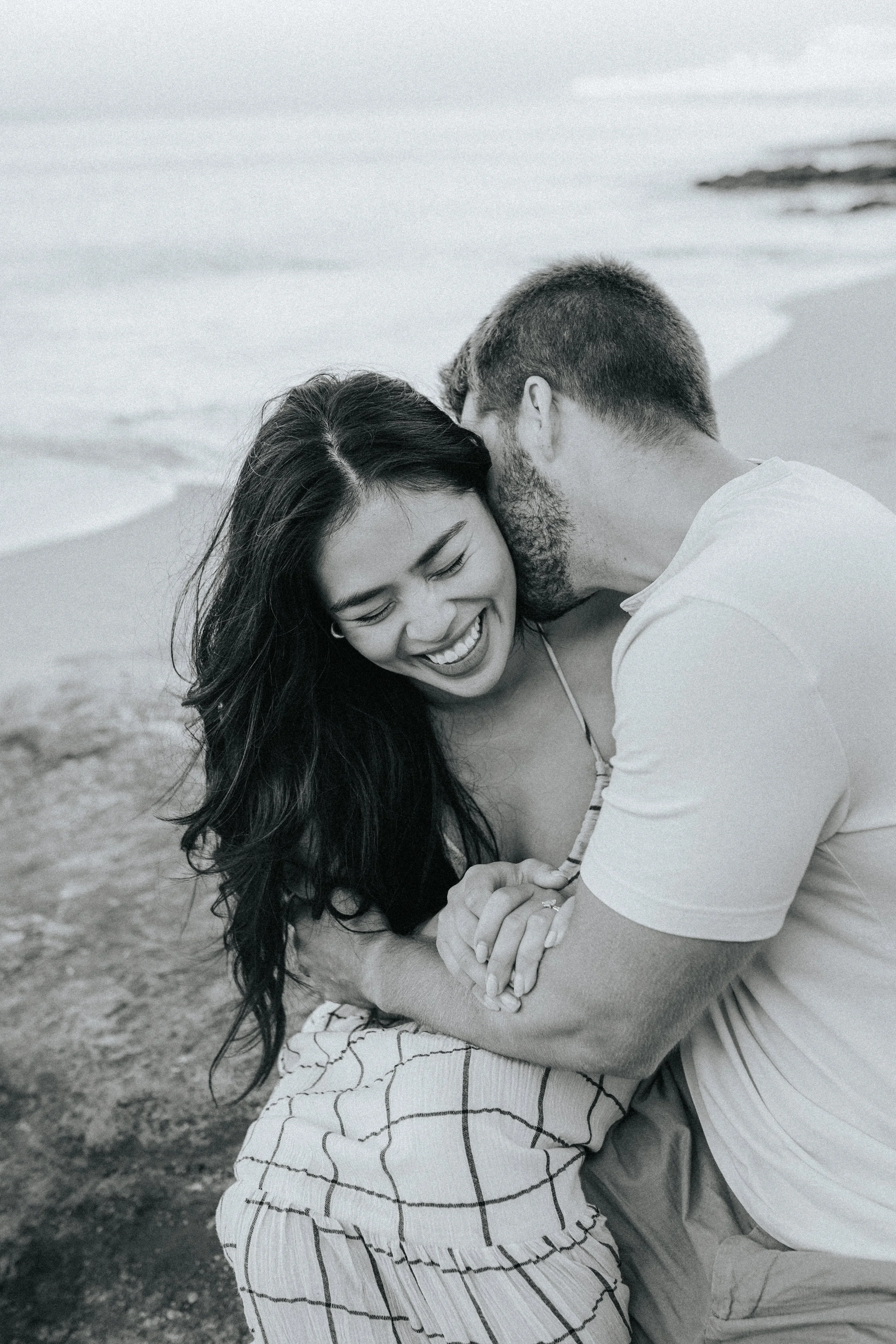 A black and white photo of a couple at the beach; the woman is smiling with her eyes closed, and the man is nuzzling her forehead, both showing affection.