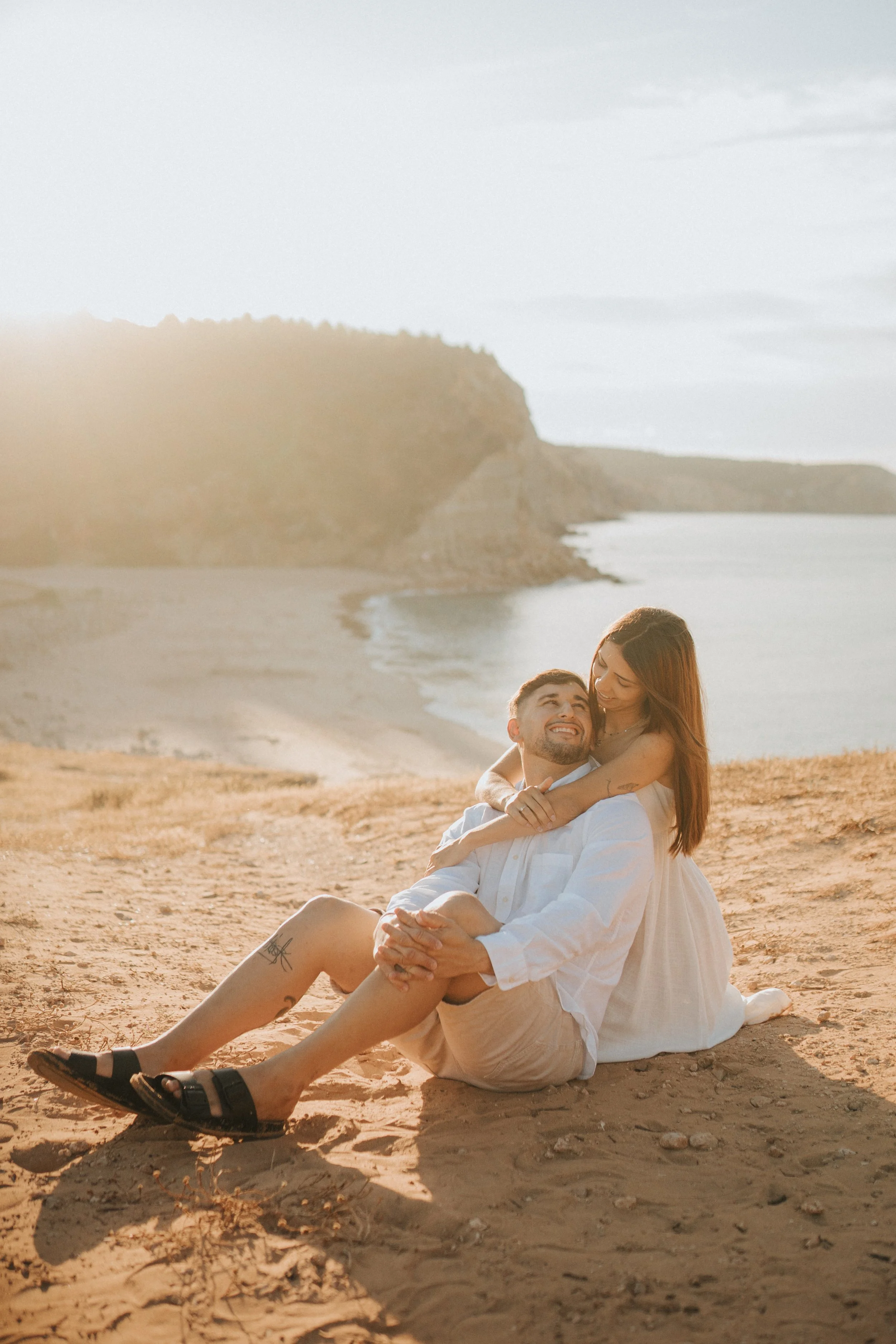 A couple sitting on the sandy beach, the woman embracing the man from behind, both smiling happily, with cliffs and the ocean in the background during sunset.