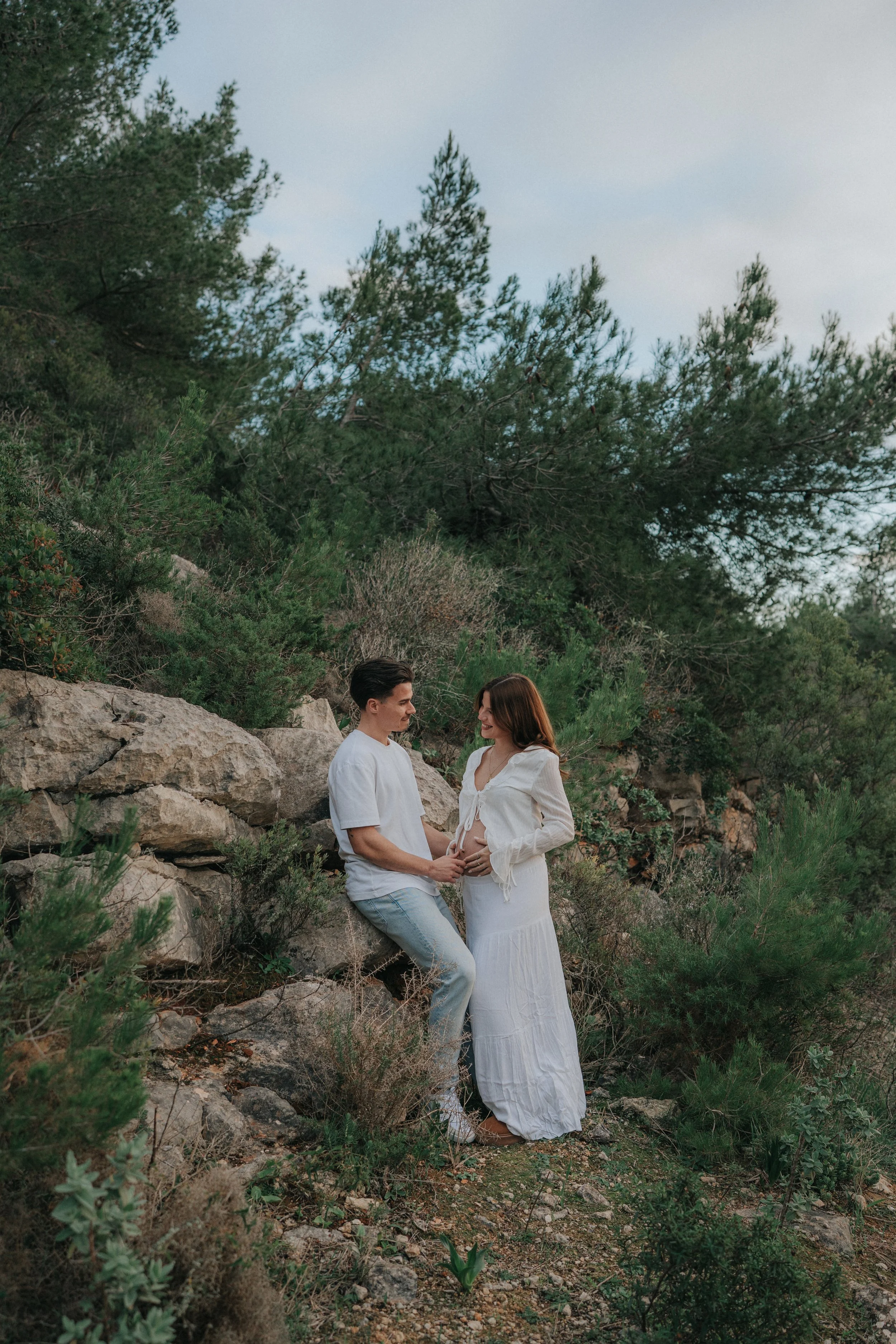 A man and pregnant woman holding hands on a rocky and lush green outdoor setting, with tall trees and a cloudy sky in the background.