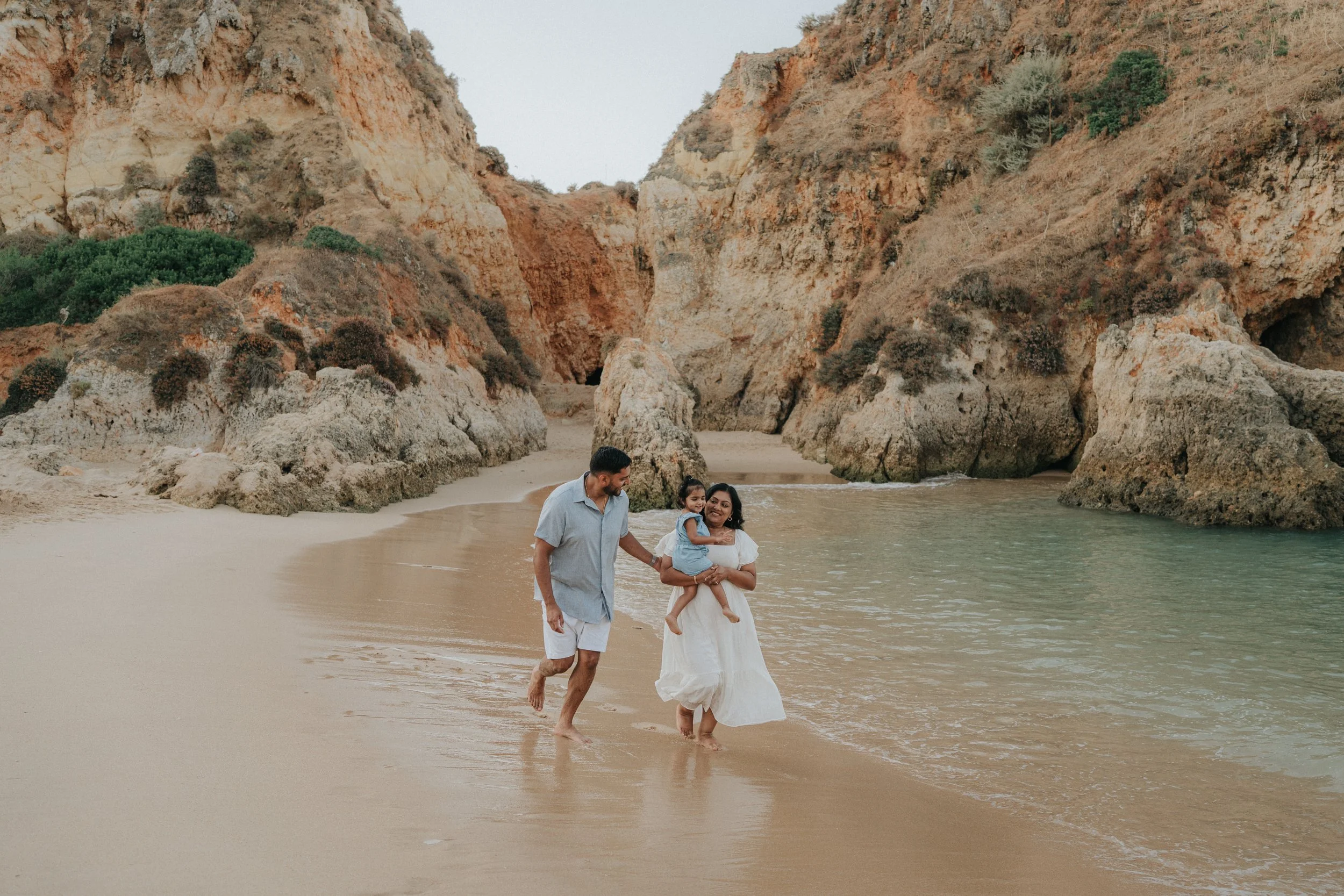 A family of three walking along the beach near the water, with rocky cliffs and sparse vegetation in the background.