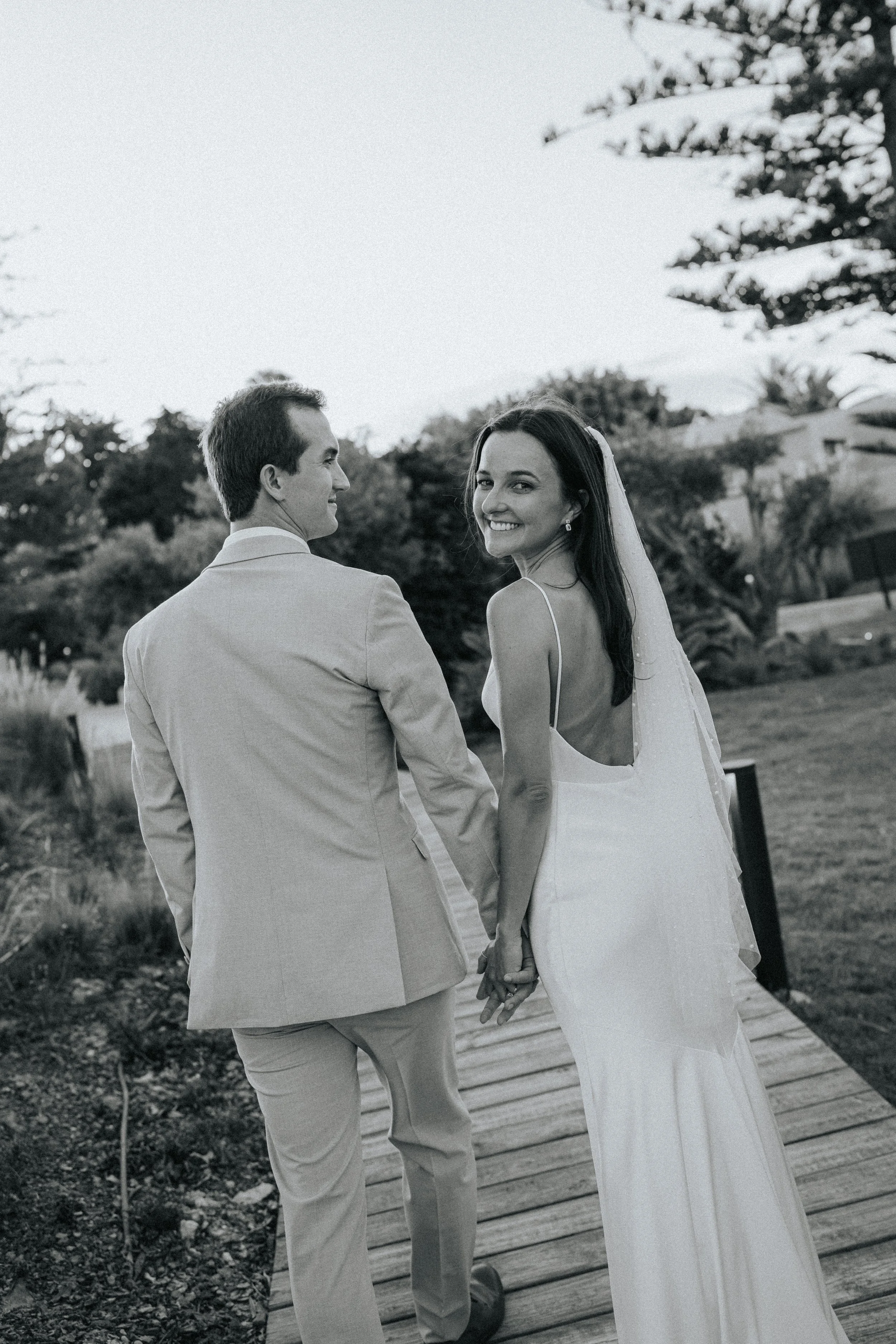A bride and groom holding hands and smiling, walking on a wooden pathway outdoors with trees and shrubs in the background.