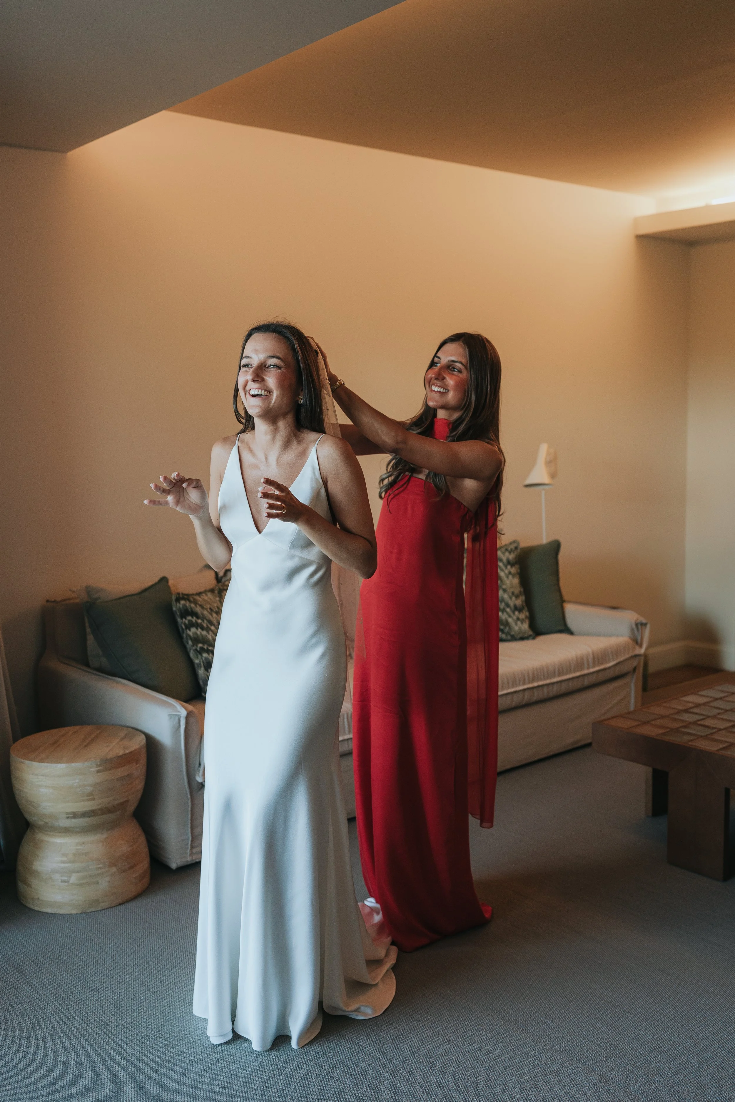 Two women in elegant dresses in a living room, one in a white gown and the other in a red gown, preparing for a formal event.