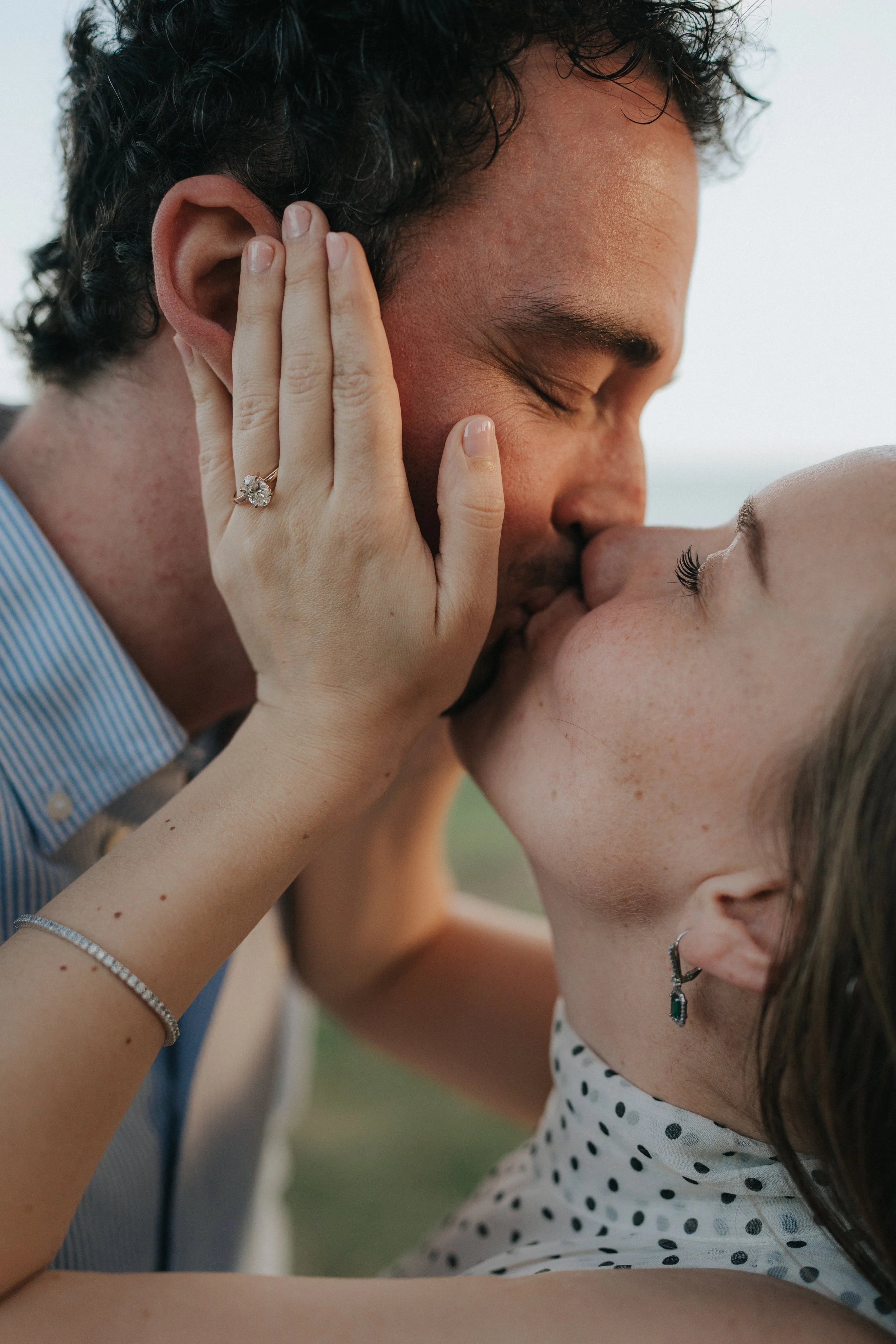 A couple sharing a kiss outdoors, close-up of their faces, with the woman's hand gently touching the man's face, showing an engagement ring, and the woman wearing a polka-dot top and earrings.