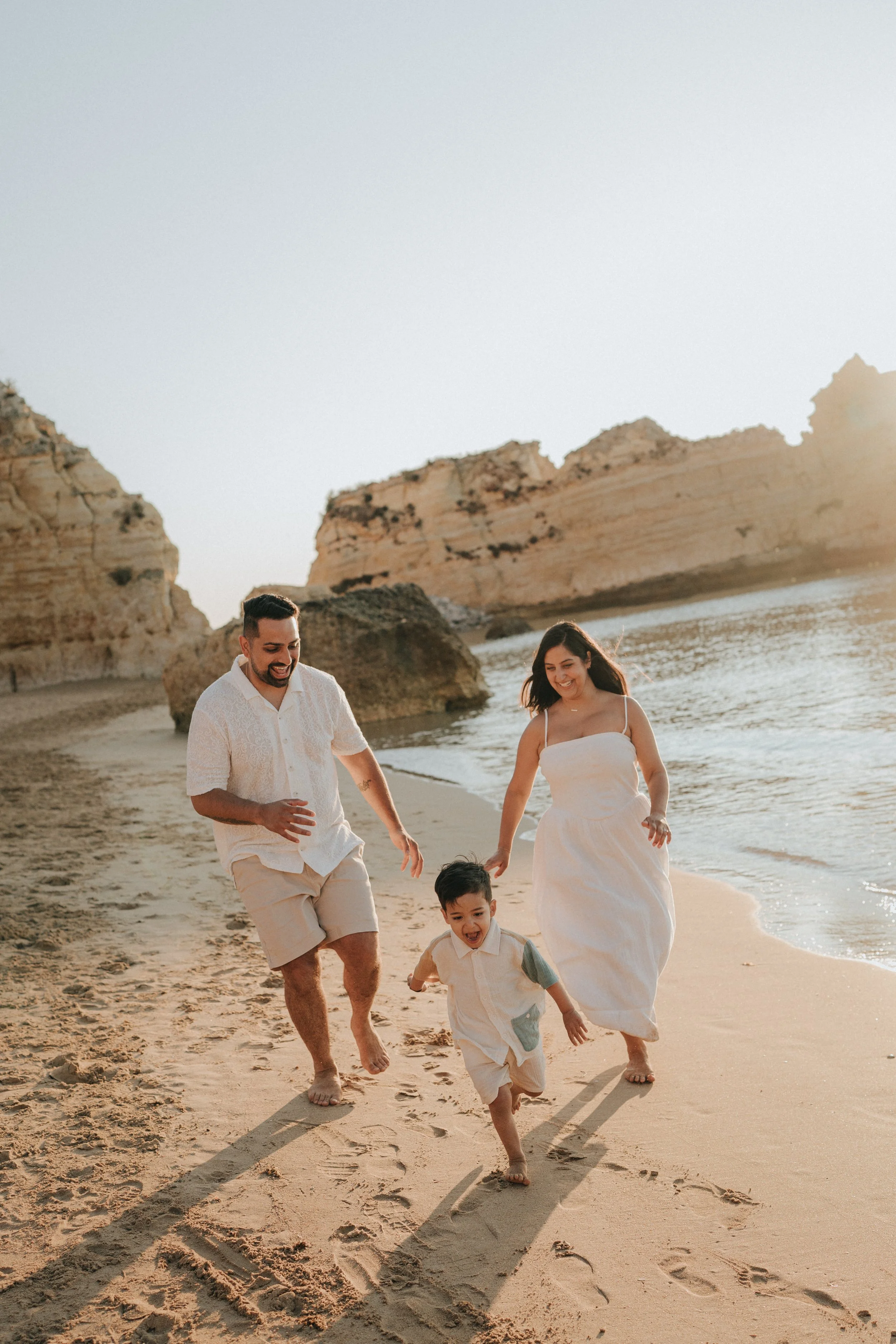 A joyful family of four, including a man, woman, and two young boys, running and playing together on a sandy beach near the ocean during sunset, with large rock formations in the background.