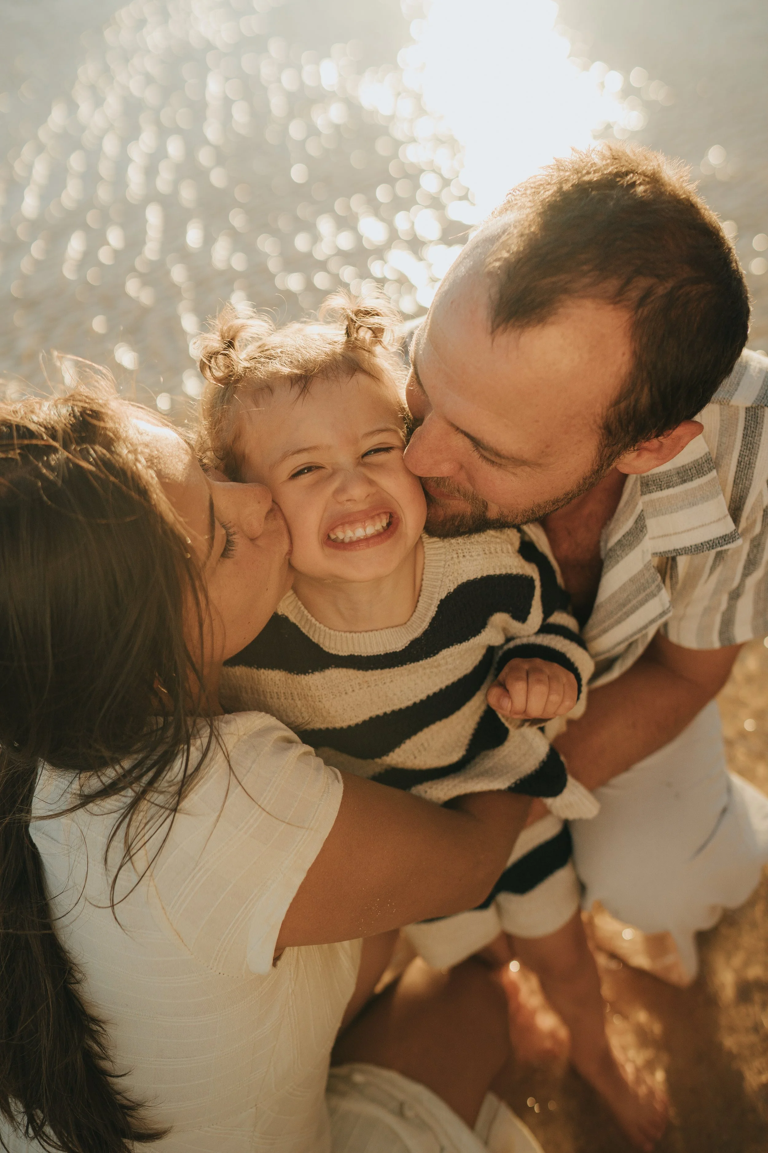 A family of three: a woman, a man, and a young girl, hugging and kissing each other outdoors near a body of water, with sunlight reflecting off the water.