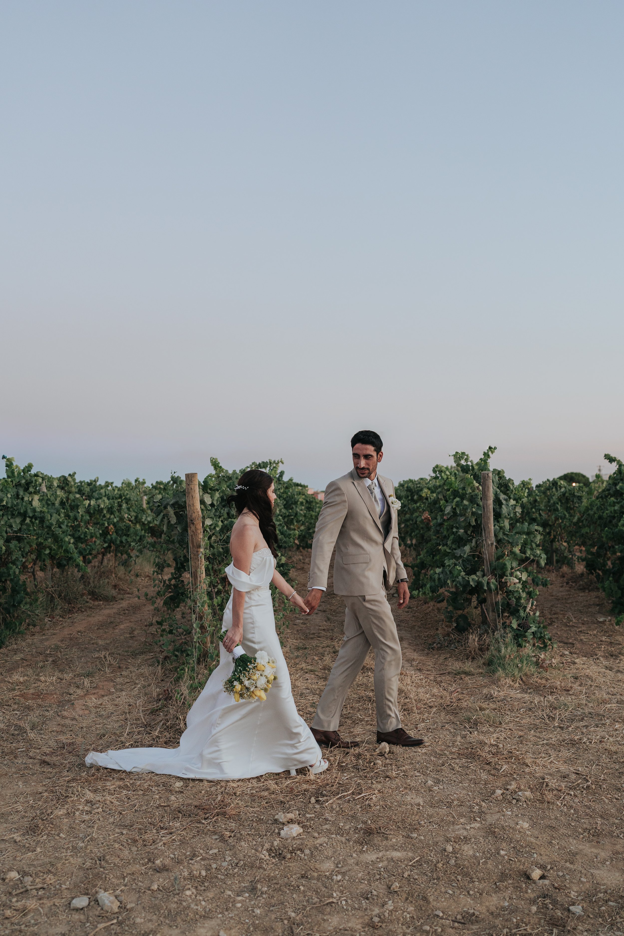 A bride and groom walking hand in hand through a vineyard during sunset. The bride is wearing a strapless white wedding dress and holding a bouquet of yellow and white flowers. The groom is in a beige suit. The sky is clear with a few clouds, and the