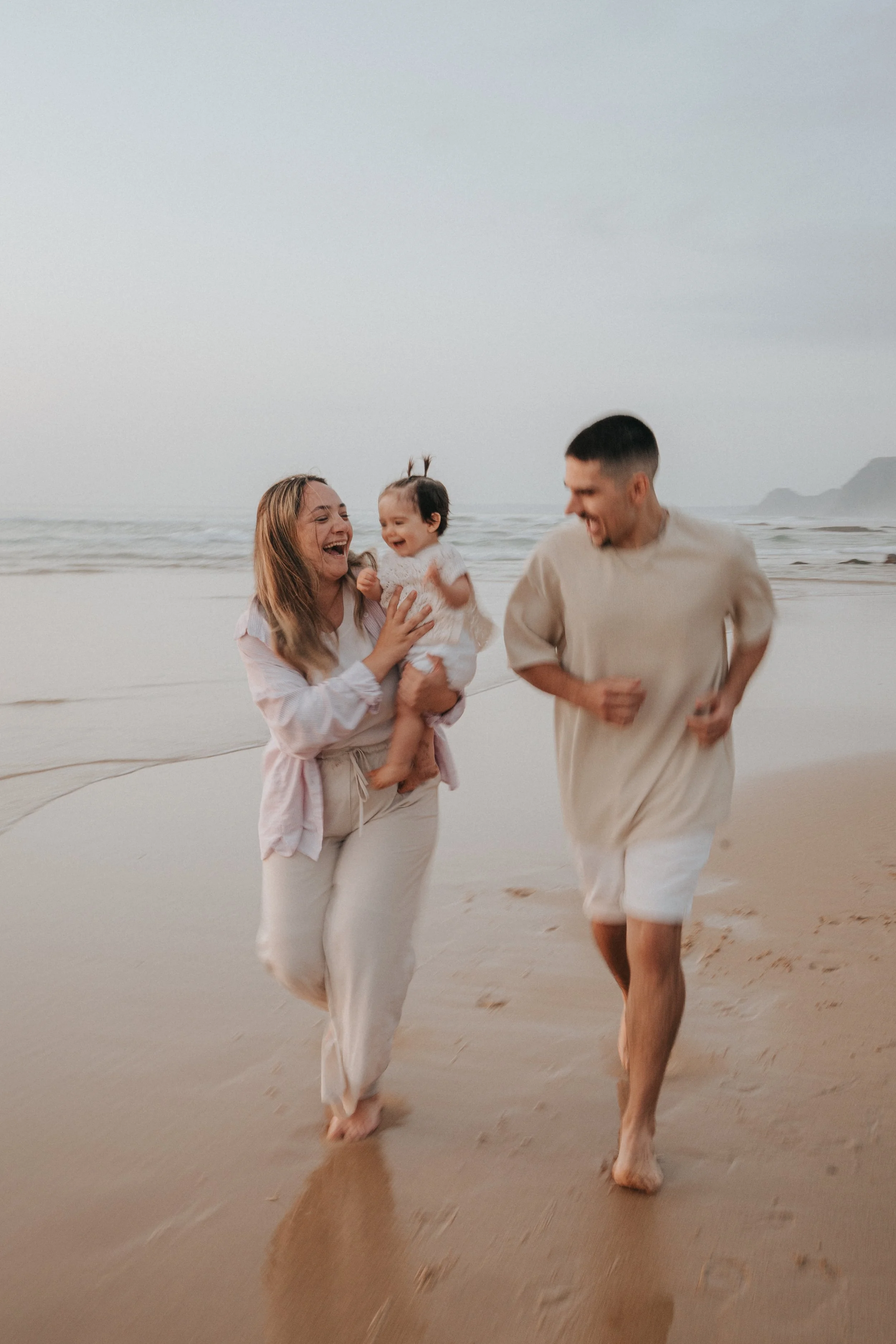 A family of three enjoying a walk on the beach with the ocean in the background, all smiling and dressed in light-colored casual clothing.
