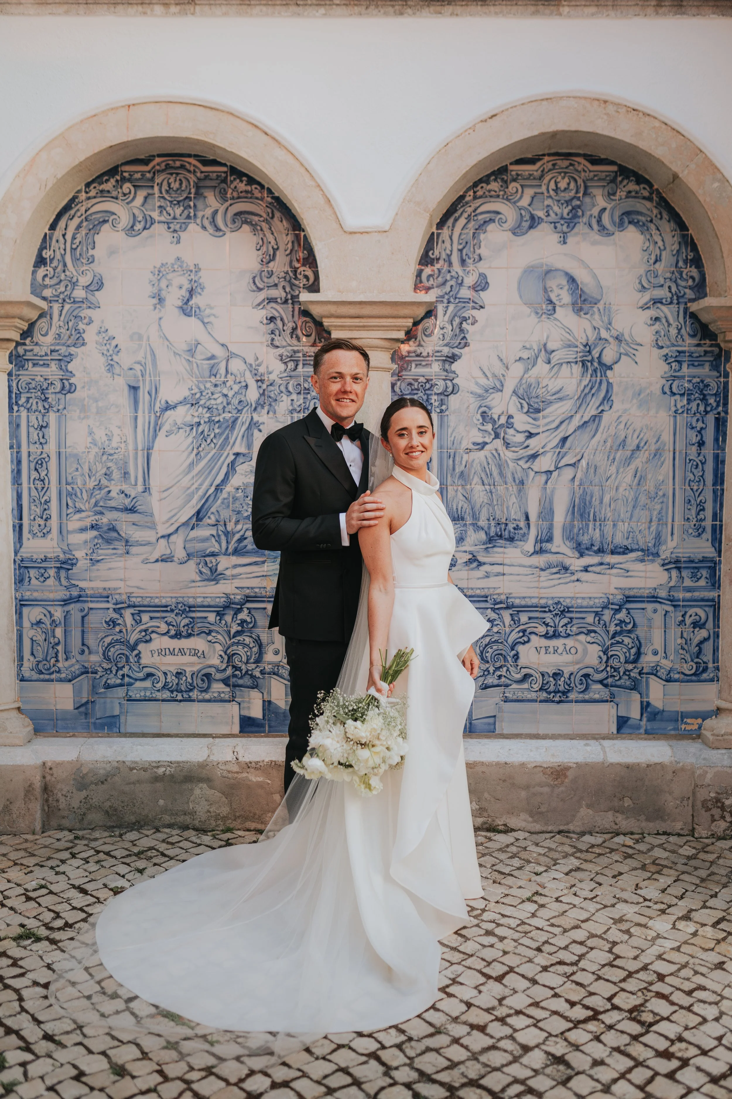 A groom in a black tuxedo and a bride in a white wedding gown standing together outdoors in front of a decorative blue and white tile mural depicting mythological or classical figures. The bride is holding a bouquet of white flowers.