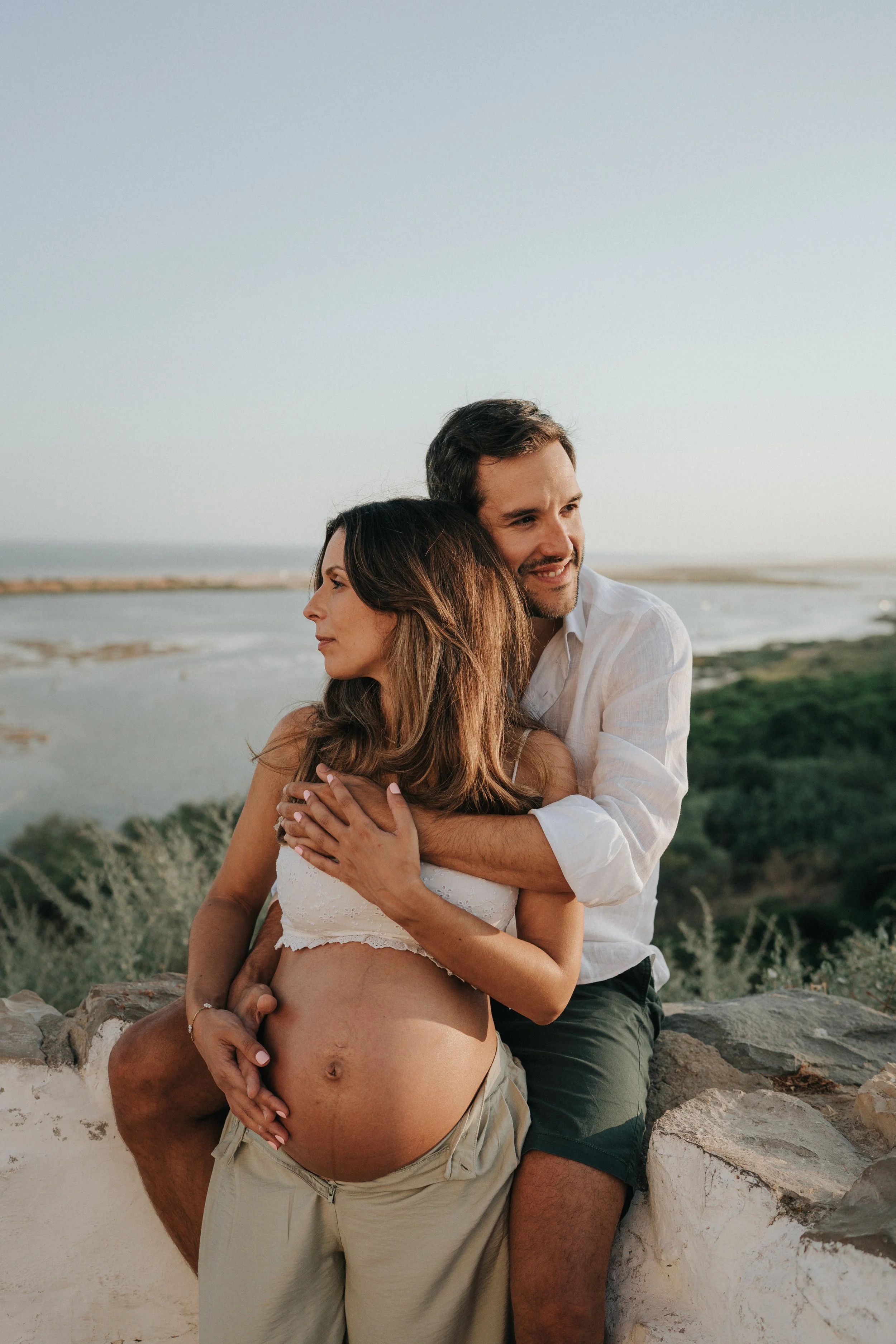A pregnant woman sitting on a rock at the beach, wearing a white bustier top, with a man behind her embracing her. They are smiling and looking to the side, with the beach and water in the background during sunset.