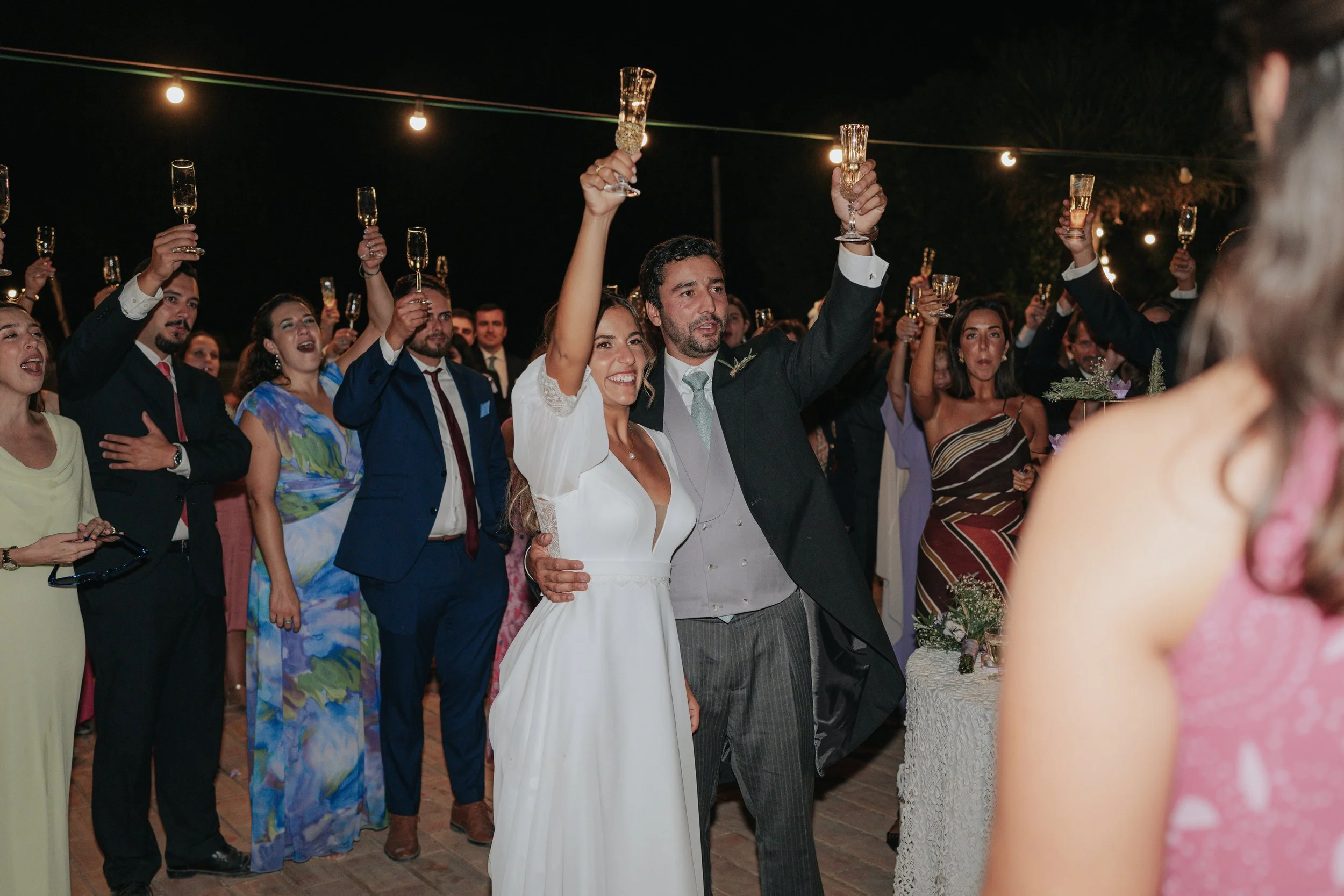 A group of people celebrating at a wedding reception, raising champagne glasses in a toast at night, outdoors decorated with string lights.