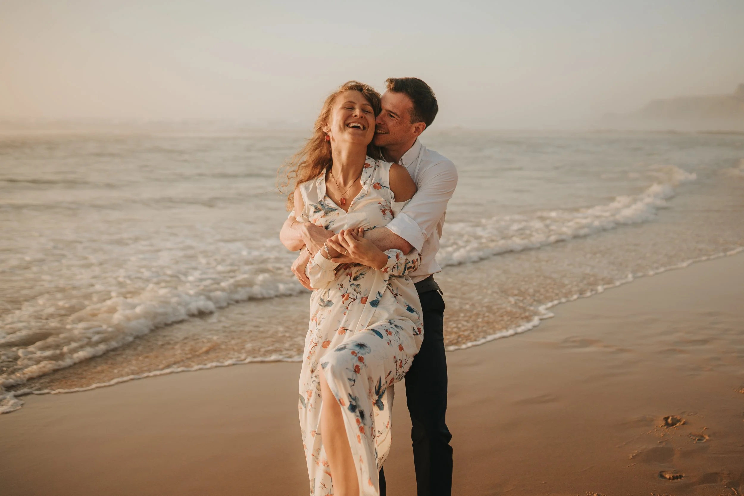 A happy couple hugging and smiling on the beach at sunset, with gentle waves in the background.