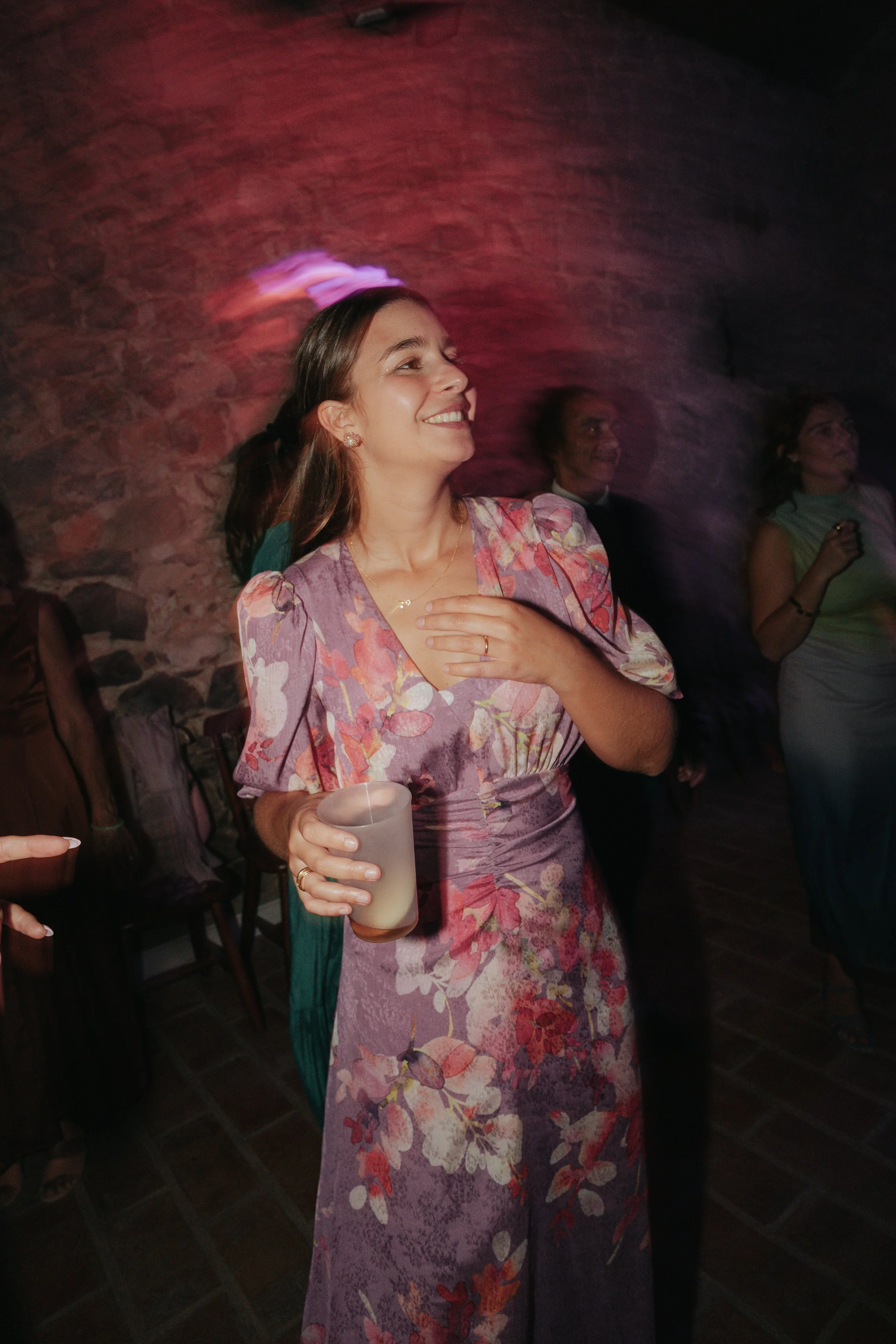 A woman in a floral dress smiling and holding a drink, with two people in the background, one man and one woman, in a dimly lit setting with a brick wall.