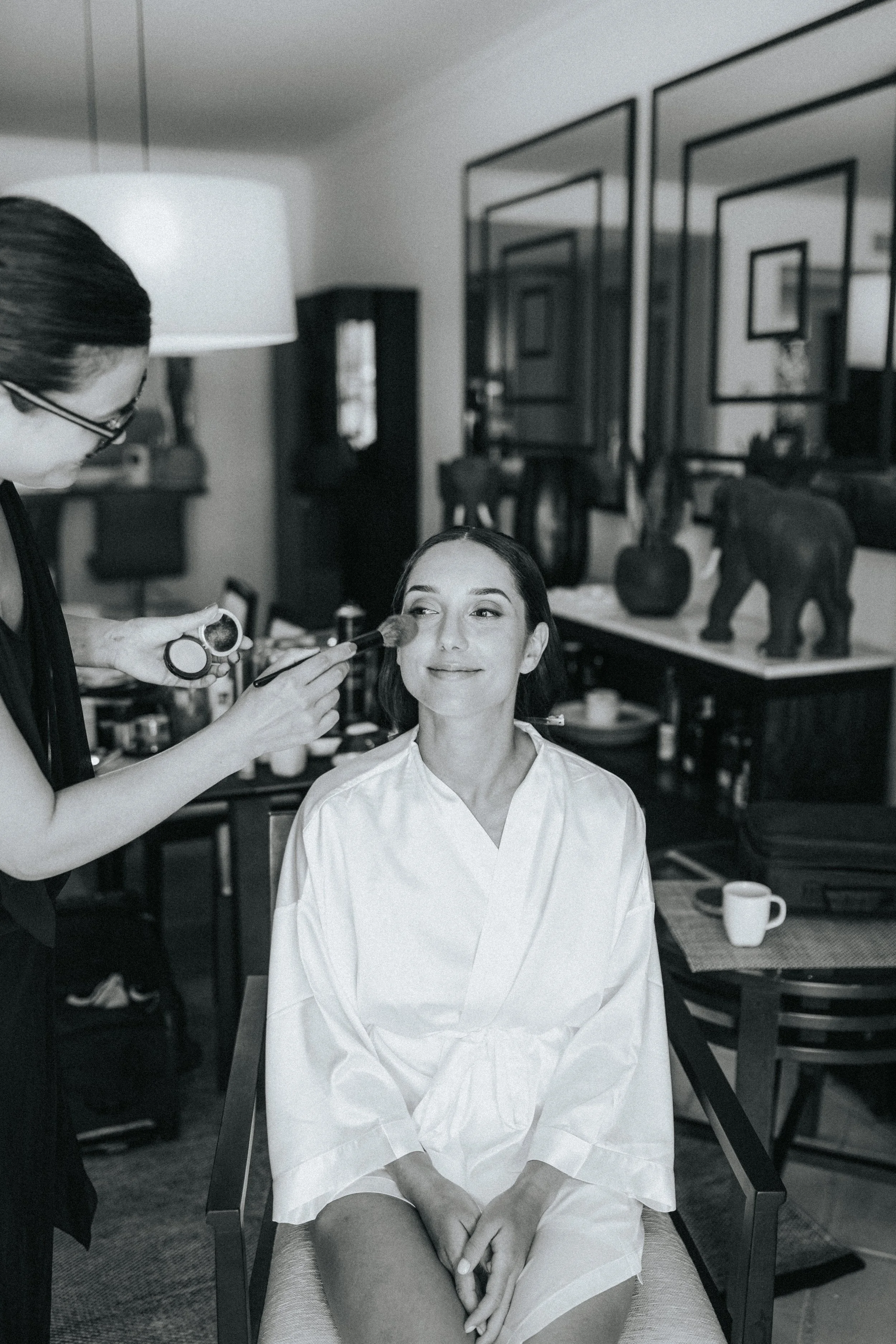 A woman is sitting in a chair with a white robe on, having her makeup applied by a makeup artist in a home setting. The makeup artist is using a brush and is wearing glasses. The background features framed artwork, a cabinet, decorative vases, and a 