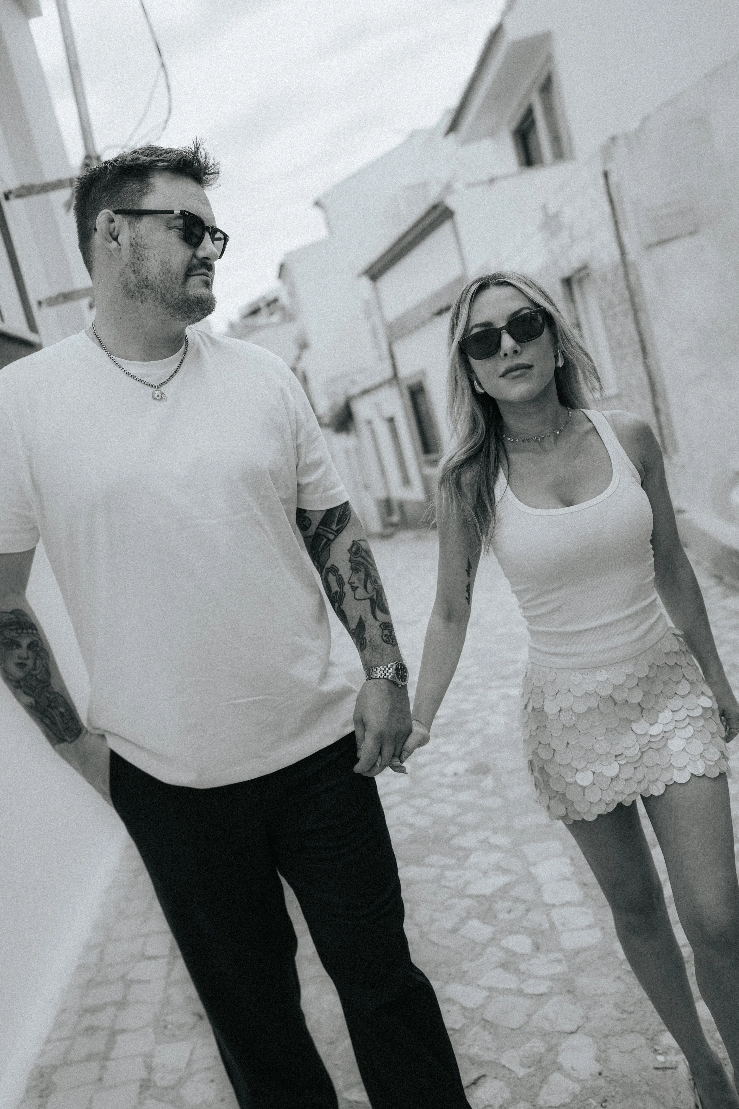 A man and woman holding hands and walking outdoors on a paved path in a sunny area. The man has short dark hair, glasses, tattoos on his arms, and is wearing a white T-shirt and a necklace. The woman has long hair, sunglasses, a necklace, and is wear