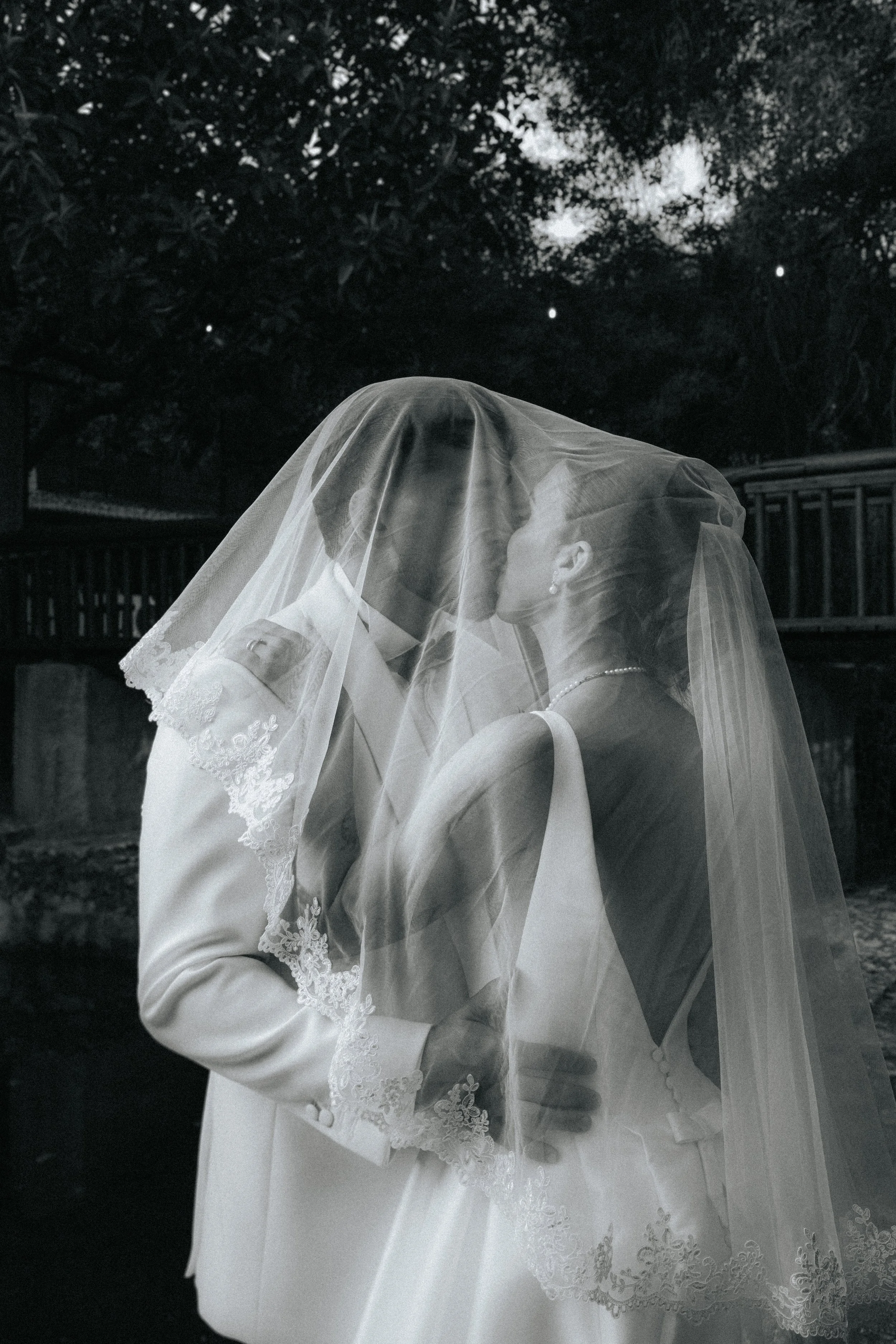 Black and white photo of a bride and groom kissing, wearing wedding attire, with a veil over the groom's face, outdoors with trees and a wooden fence in the background.