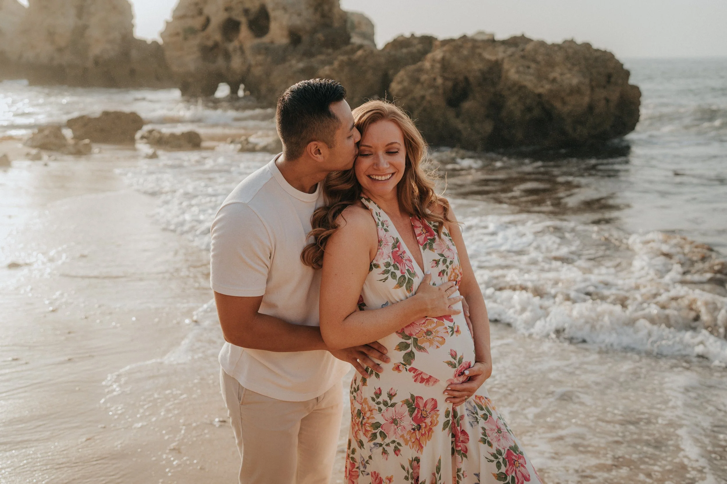 A couple standing on the beach, with the man kissing the woman's forehead. The woman is pregnant, wearing a floral dress, and smiling. The scene is set by the ocean with rocks in the background during sunset.