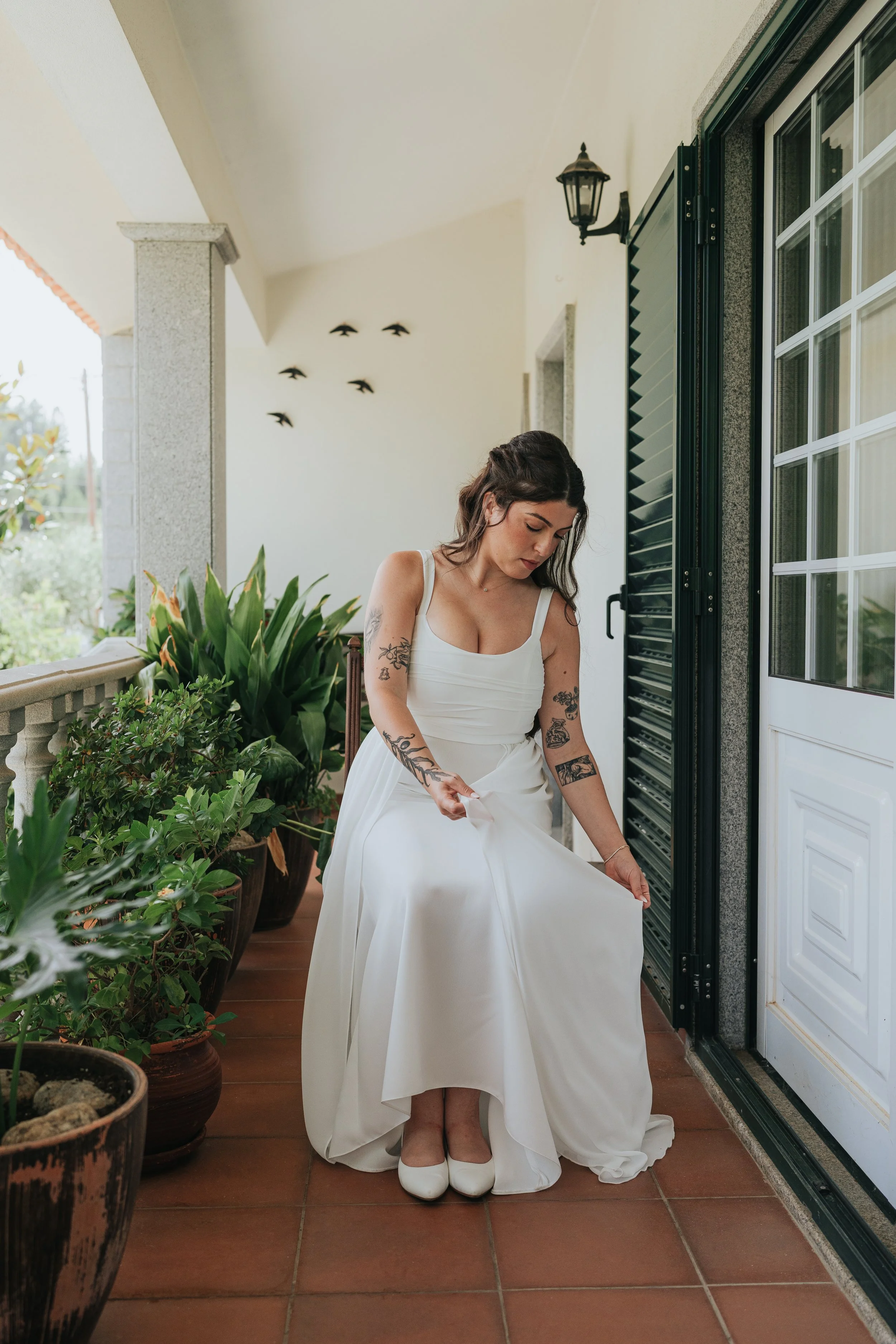 A woman in a white dress is sitting on a porch, adjusting her dress. She has tattoos on her arms and is wearing white shoes. There are potted plants on the porch, and a window with black shutters behind her. The porch has a tiled floor and a white wa