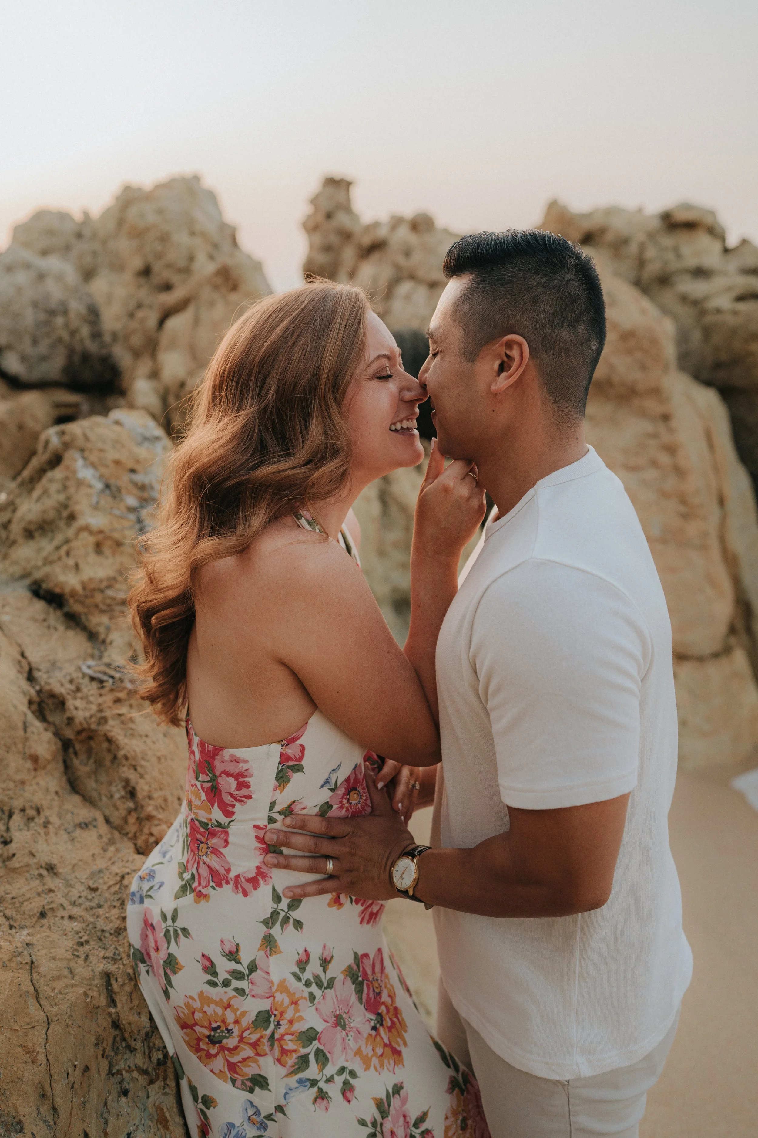 A couple standing closely together, smiling and touching noses in front of a rocky background during sunset or dusk.