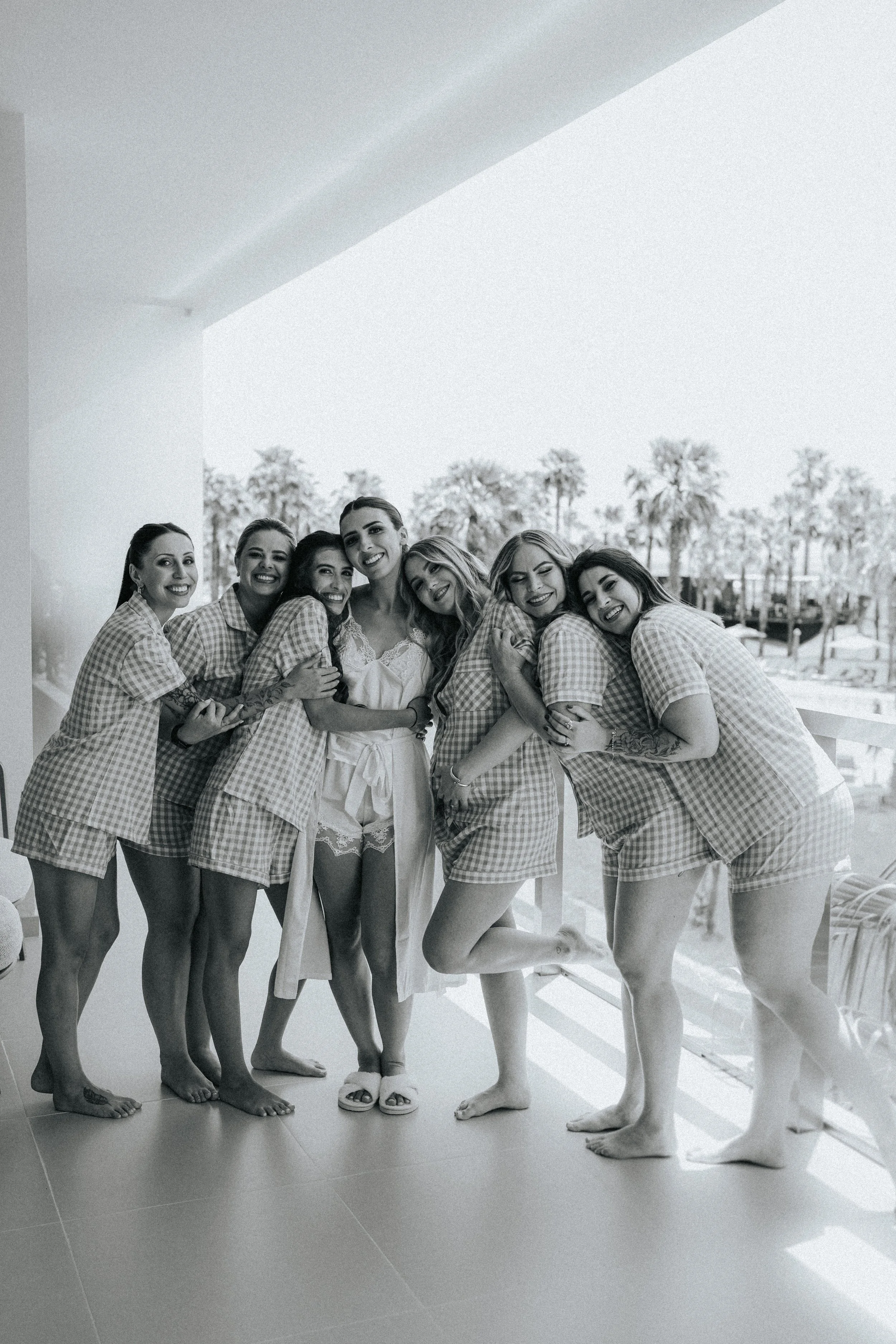 Group of women wearing matching checkered nightwear, smiling and embracing each other outdoors with trees and a walkway in the background.