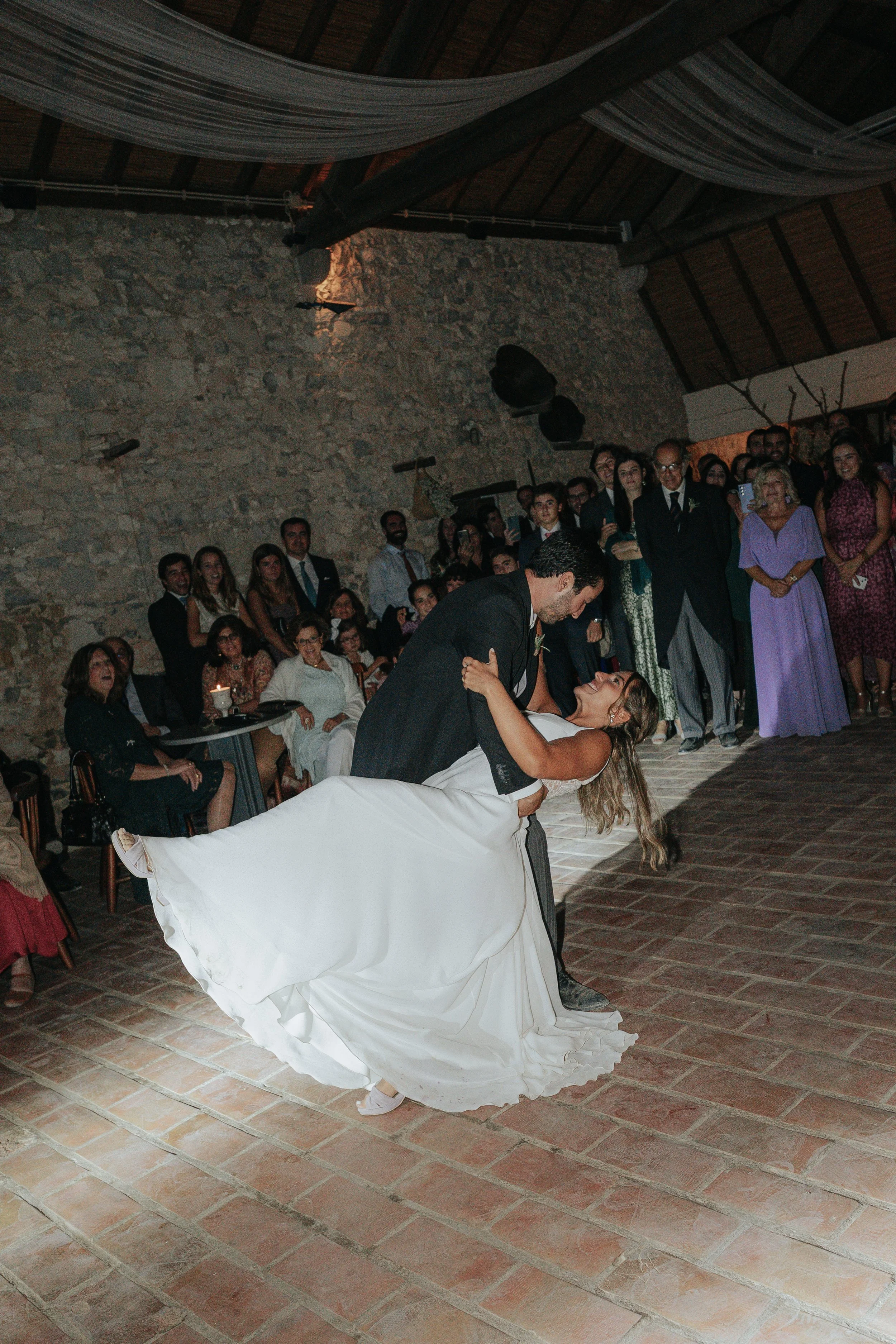 A groom dips and kisses the bride during their wedding dance in front of an audience inside a rustic venue with stone walls and wooden ceiling beams.
