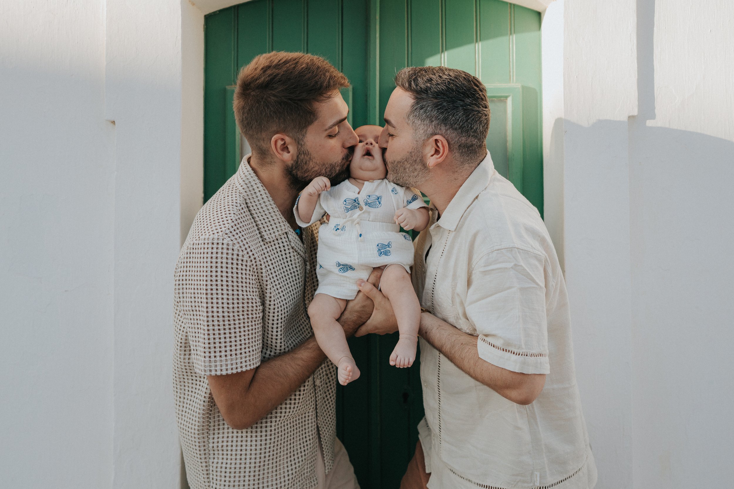 Two men kissing a baby on each cheek, holding the baby between them. The men are smiling with closed eyes, the baby looks at one of the men, wearing a white shirt with blue fish print, standing in front of a green door with shadows on a white wall.