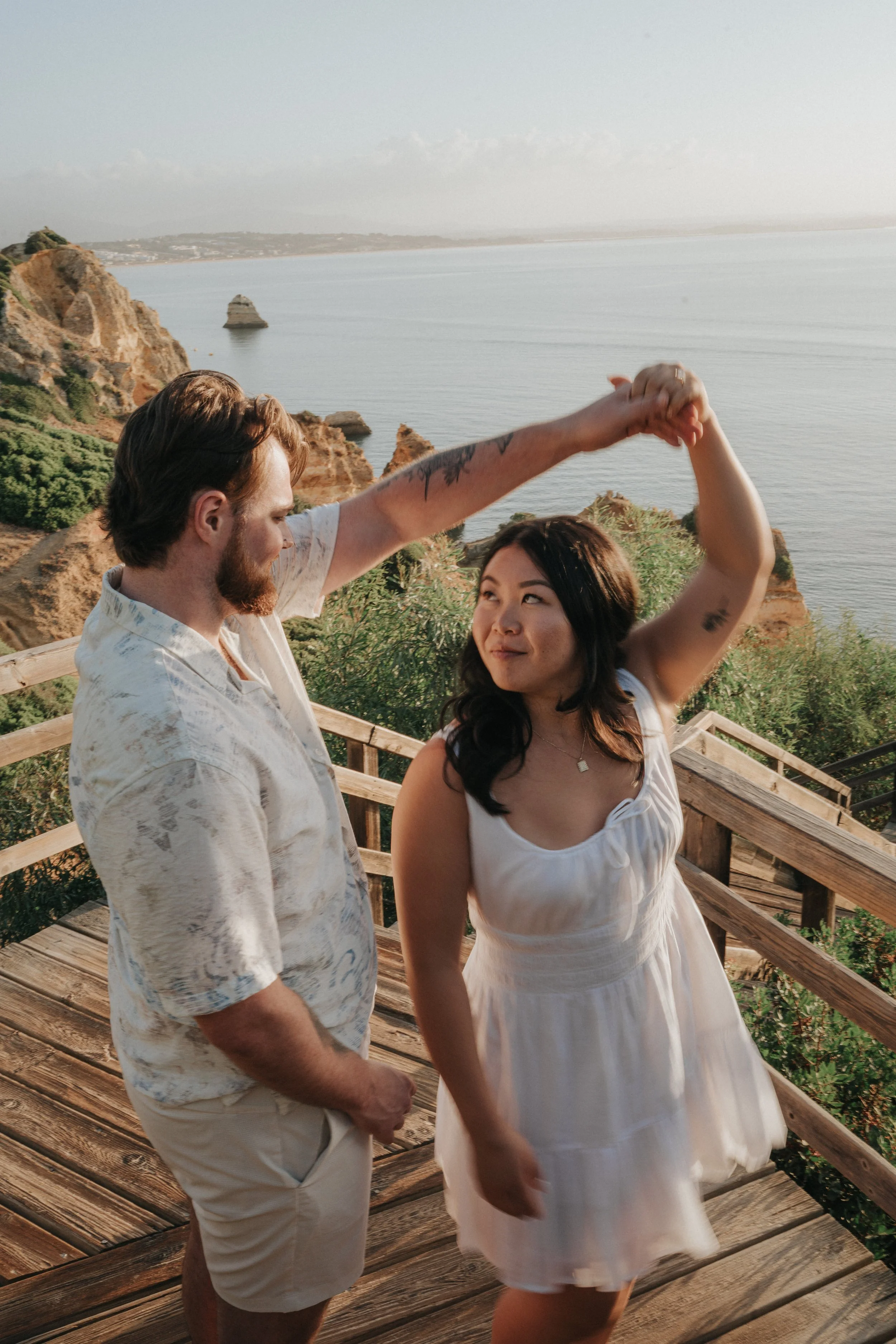 A man and woman are dancing on a wooden deck overlooking the ocean and cliffs, holding hands and smiling at each other.
