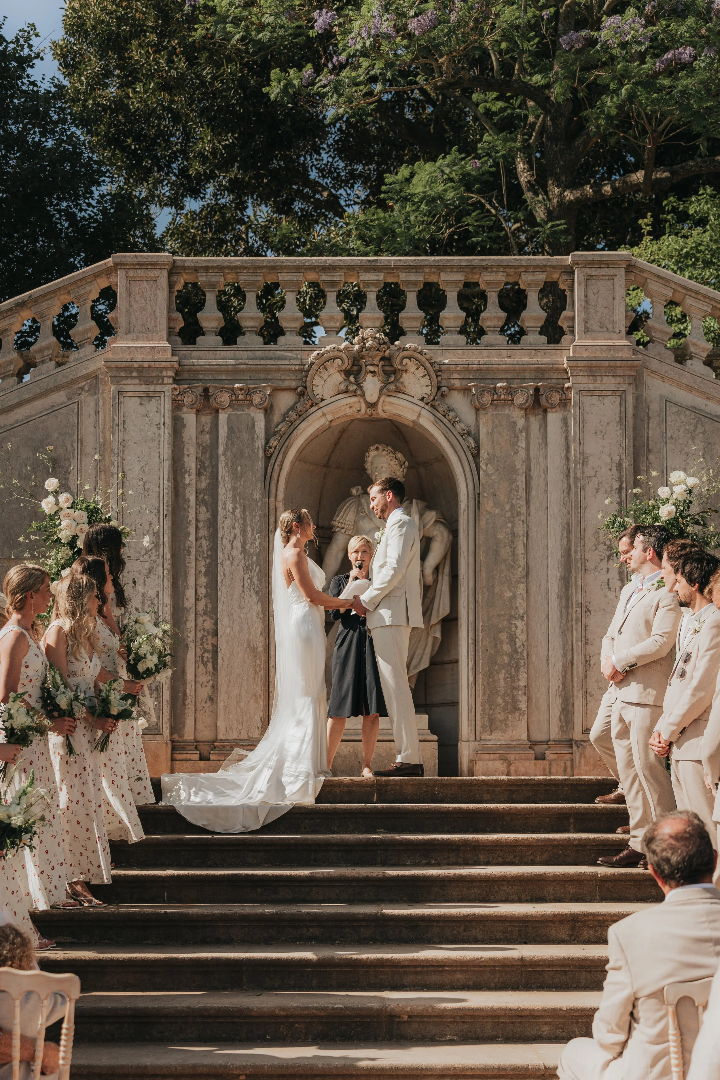 A couple exchanges vows at an outdoor wedding altar with guests witnessing, floral arrangements, and a historic stone building with statues and decorative stonework in the background.