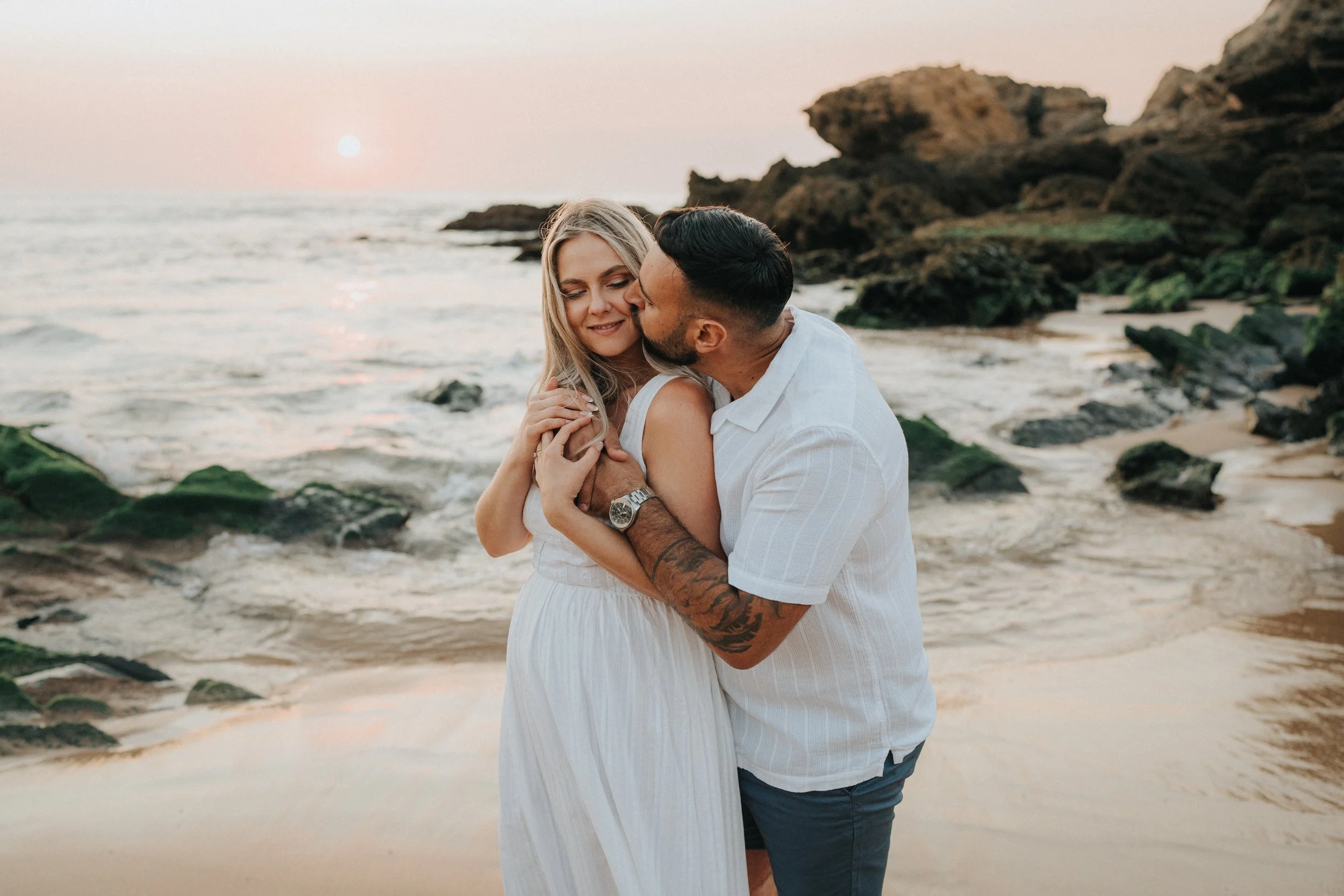 A romantic scene of a couple embracing on a beach at sunset, with rocks and the ocean in the background.