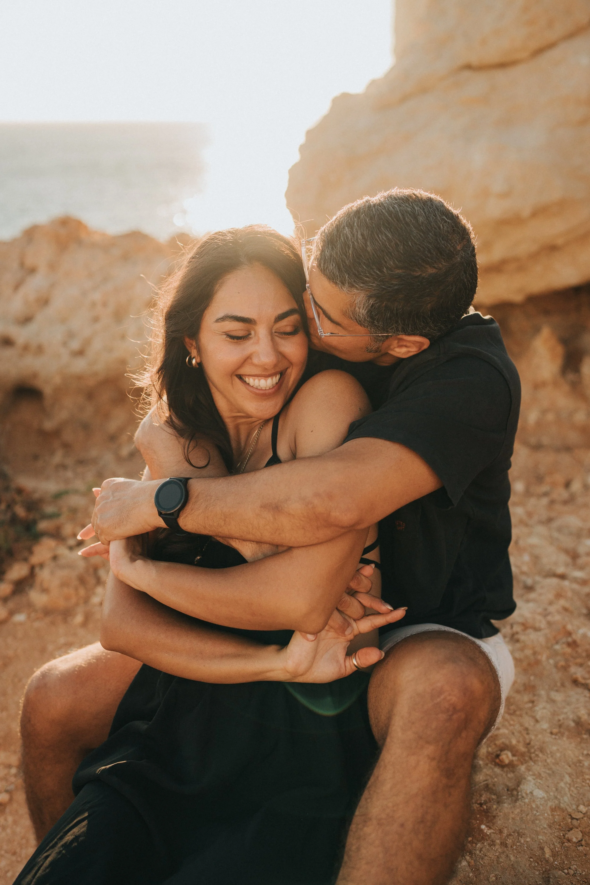 A man and a woman hugging and smiling on a sandy beach with rocks in the background during sunset.