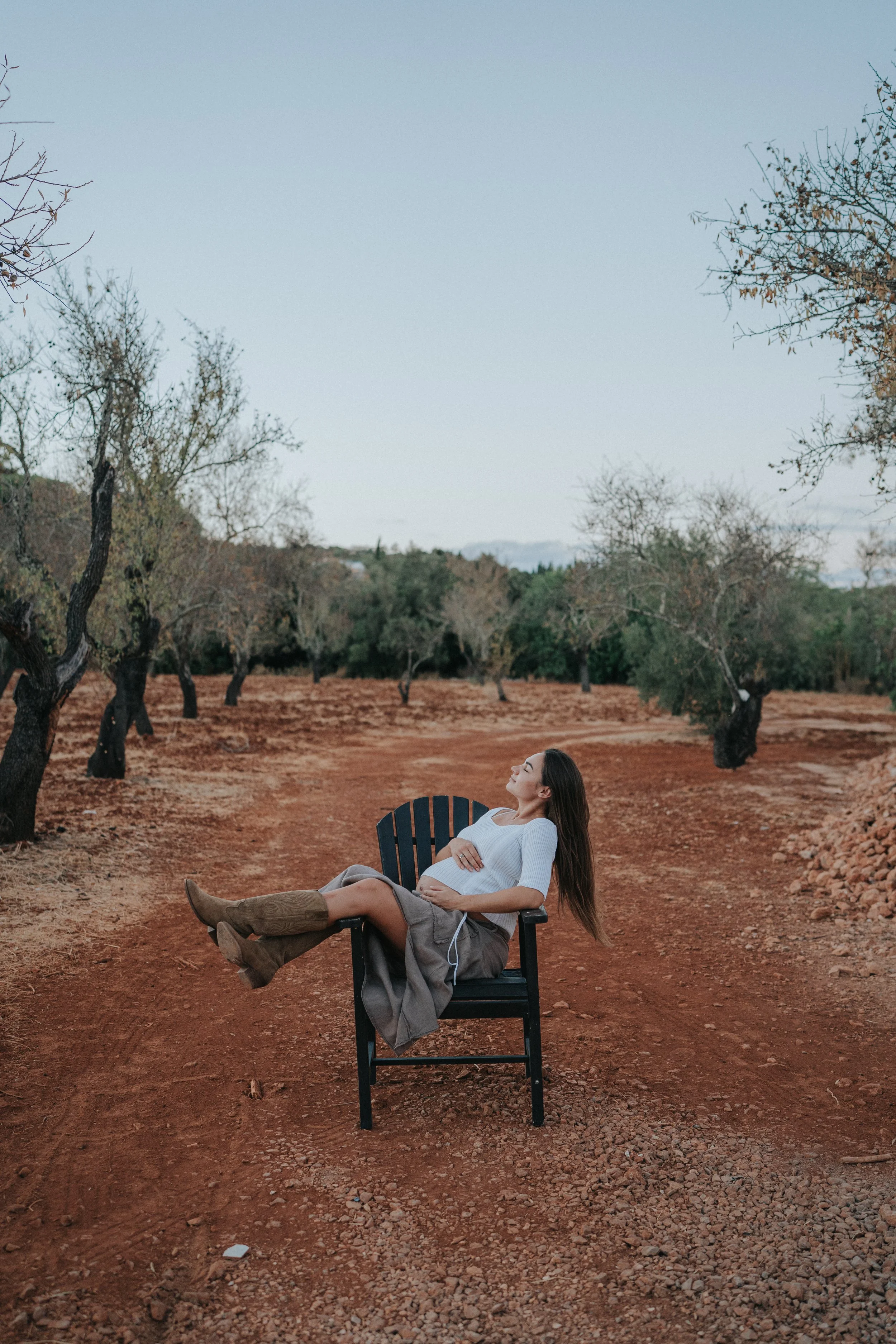 A pregnant woman with long brown hair and a white top sitting relaxed on a black chair outdoors, surrounded by leafless trees and dry soil, with a clear sky overhead.