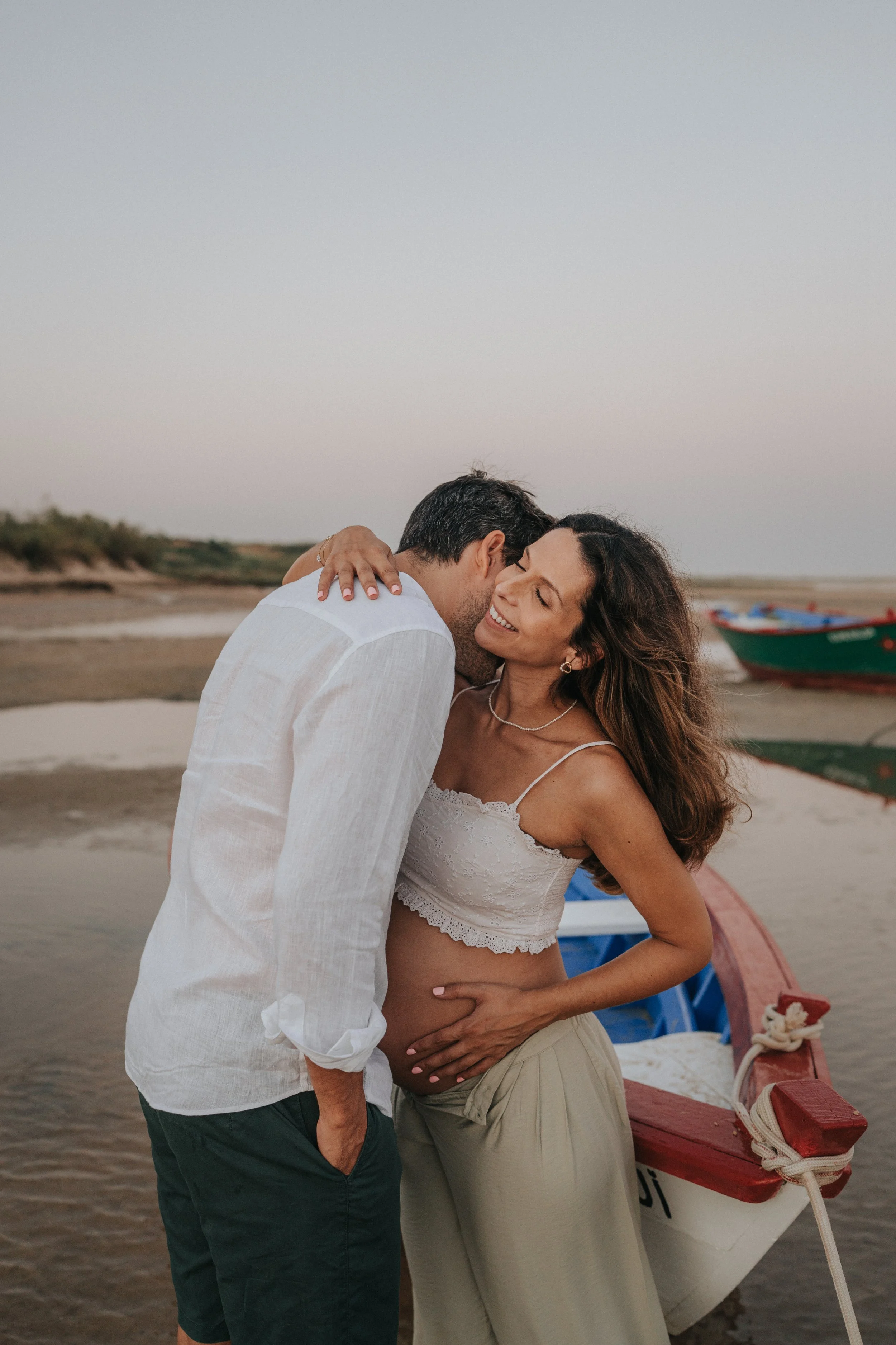 A man and a woman embrace on a beach during sunset, with a small boat tied to a dock in the background.