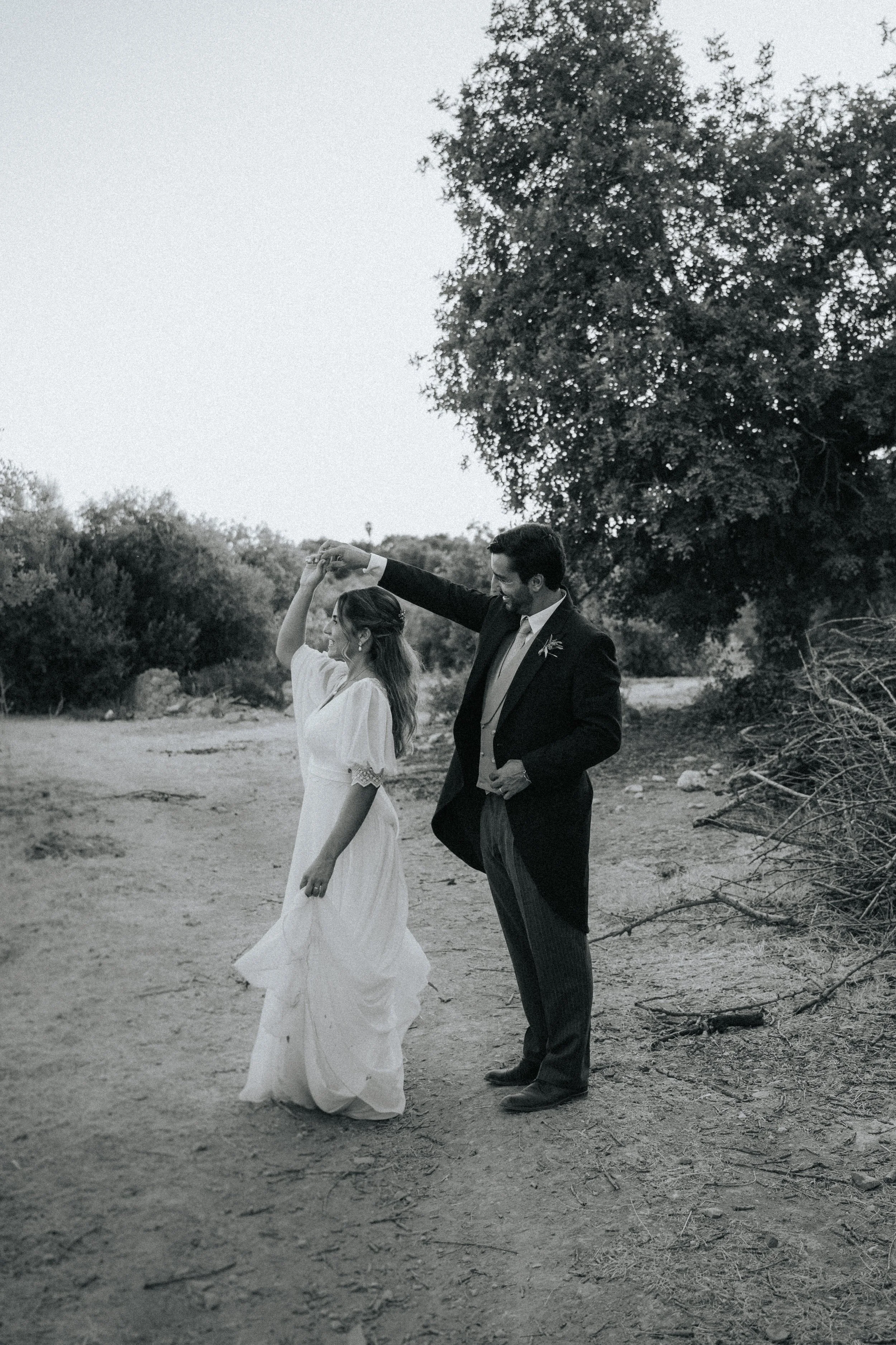 A black-and-white photo of a couple dancing outdoors, with the man in a suit and the woman in a flowing dress, holding hands and smiling. They are surrounded by trees and natural landscape.