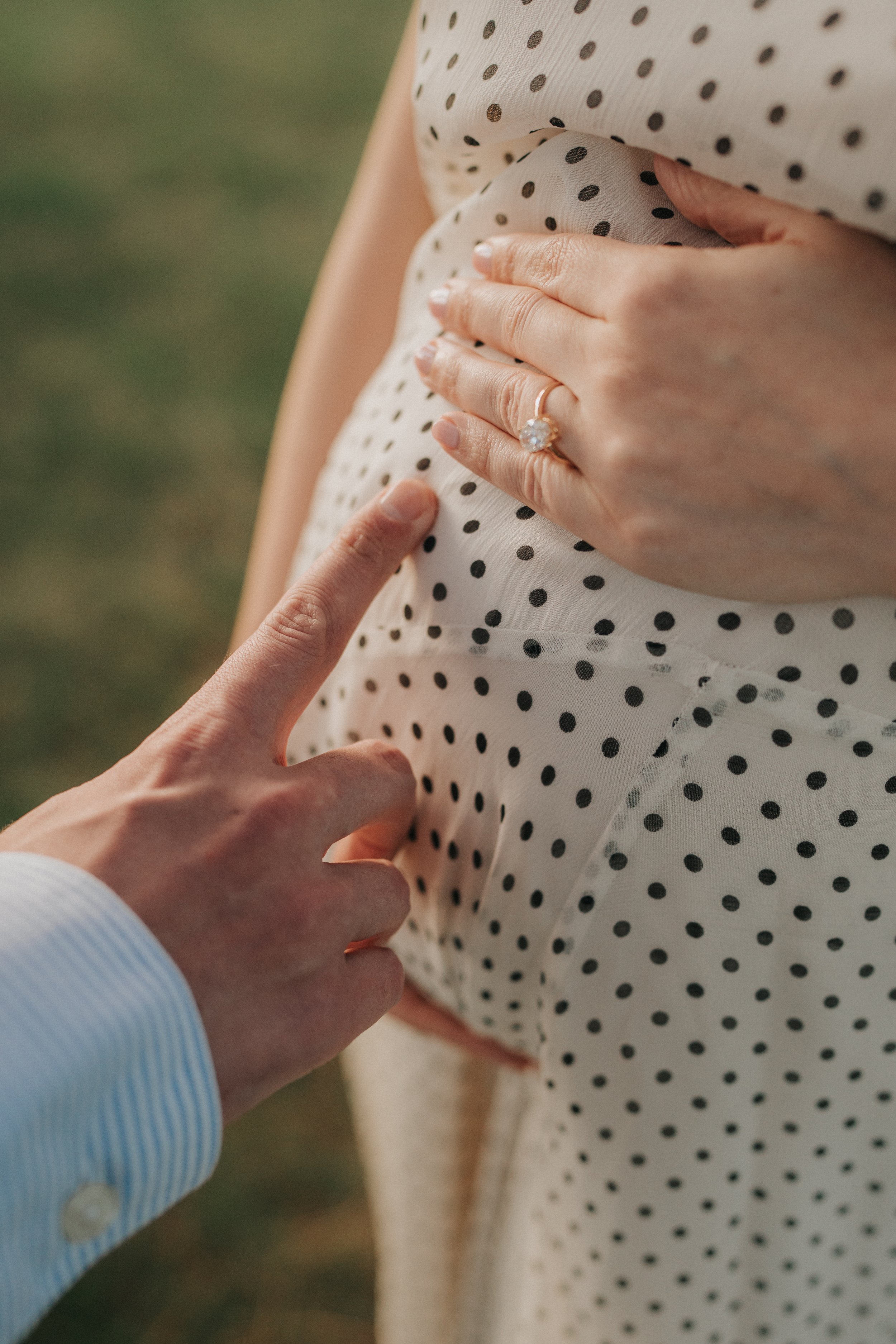 Close-up of a woman's hand placing a ring on her finger, wearing a polka dot dress, outdoor setting.