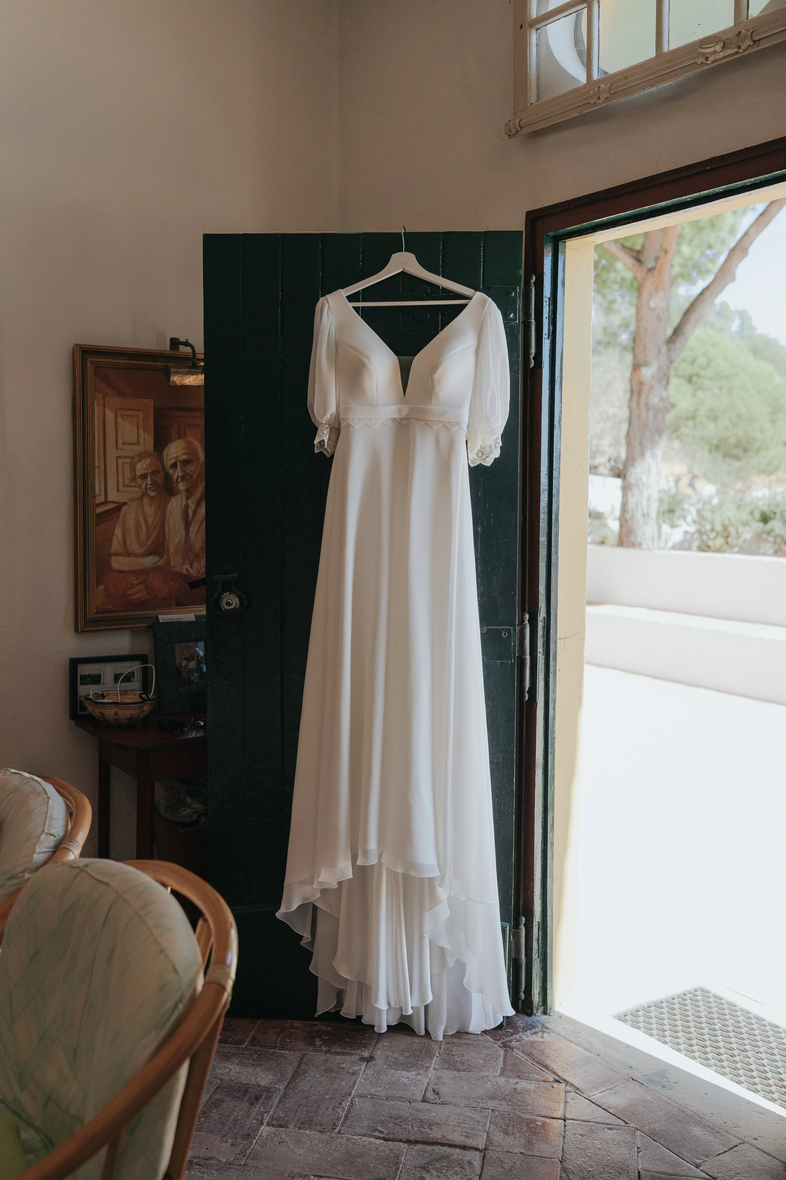 A white wedding dress with puffy long sleeves and lace cuffs hanging on a hanger in a room at an open door with sunlight and trees outside.