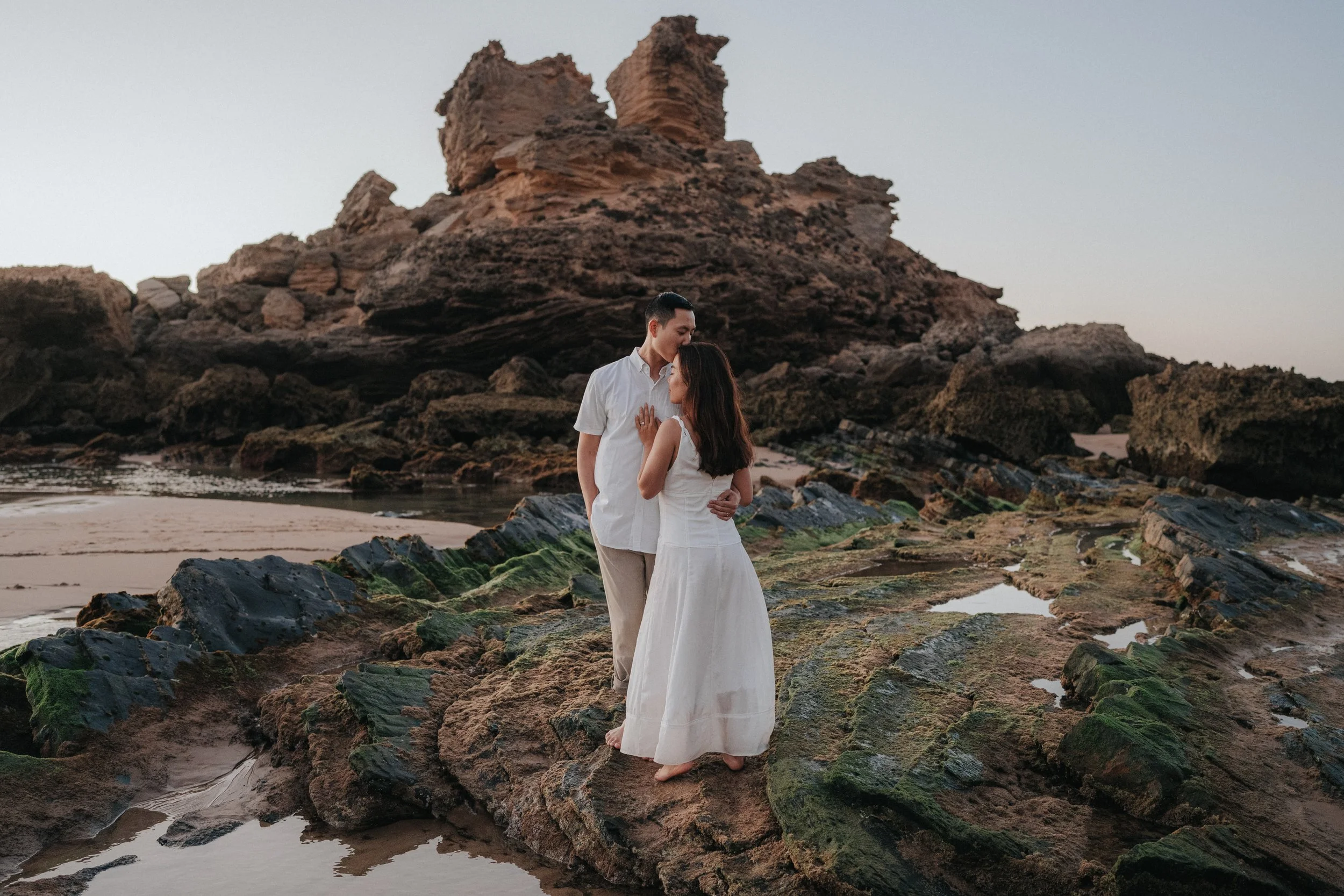 A couple standing closely together on a rocky beach with large rock formations in the background, during sunset or dusk, with calm water surrounding them.