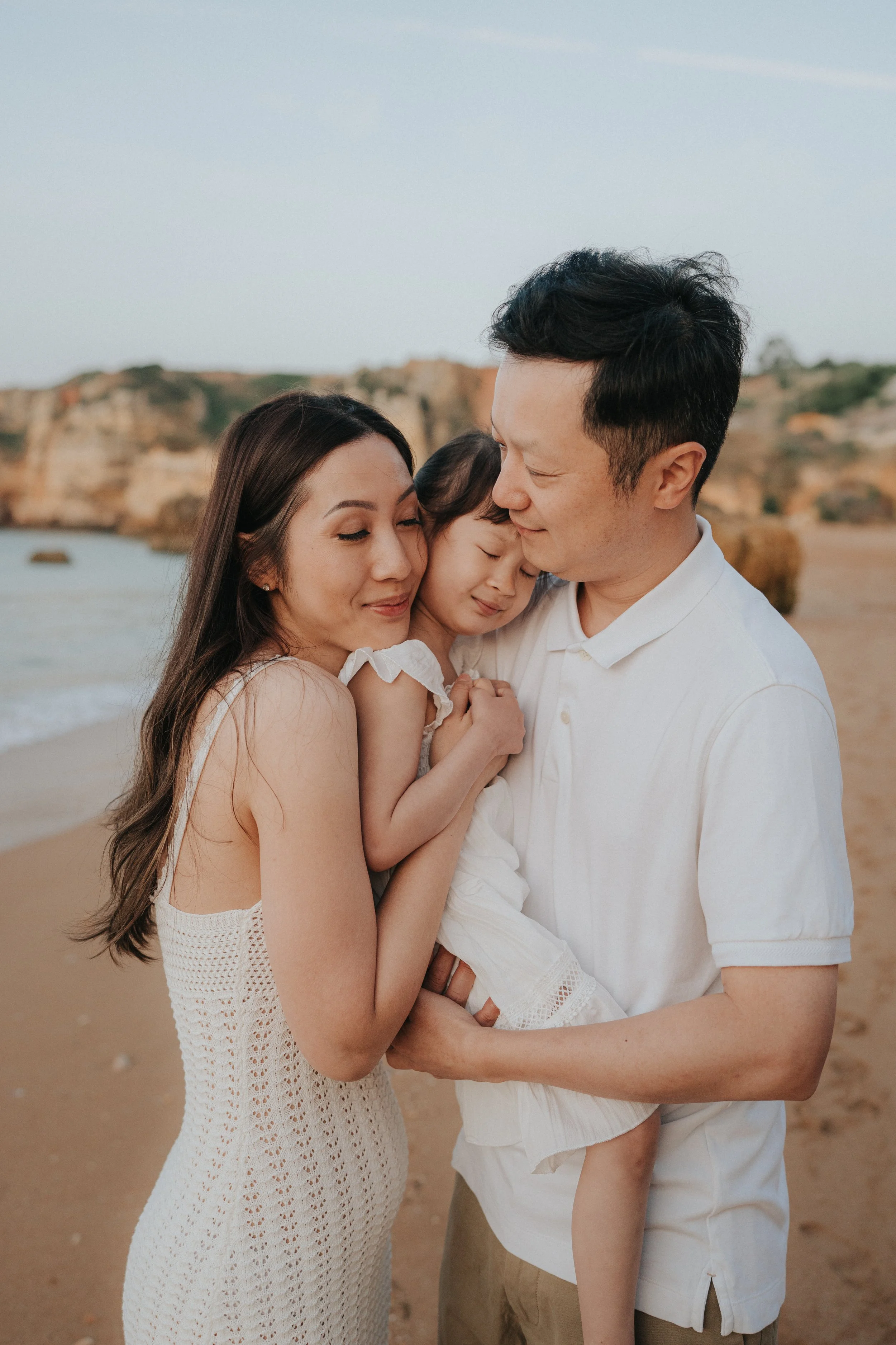 A family of three, a mother, father, and young daughter, hugging on a sandy beach with cliffs in the background, during sunset.