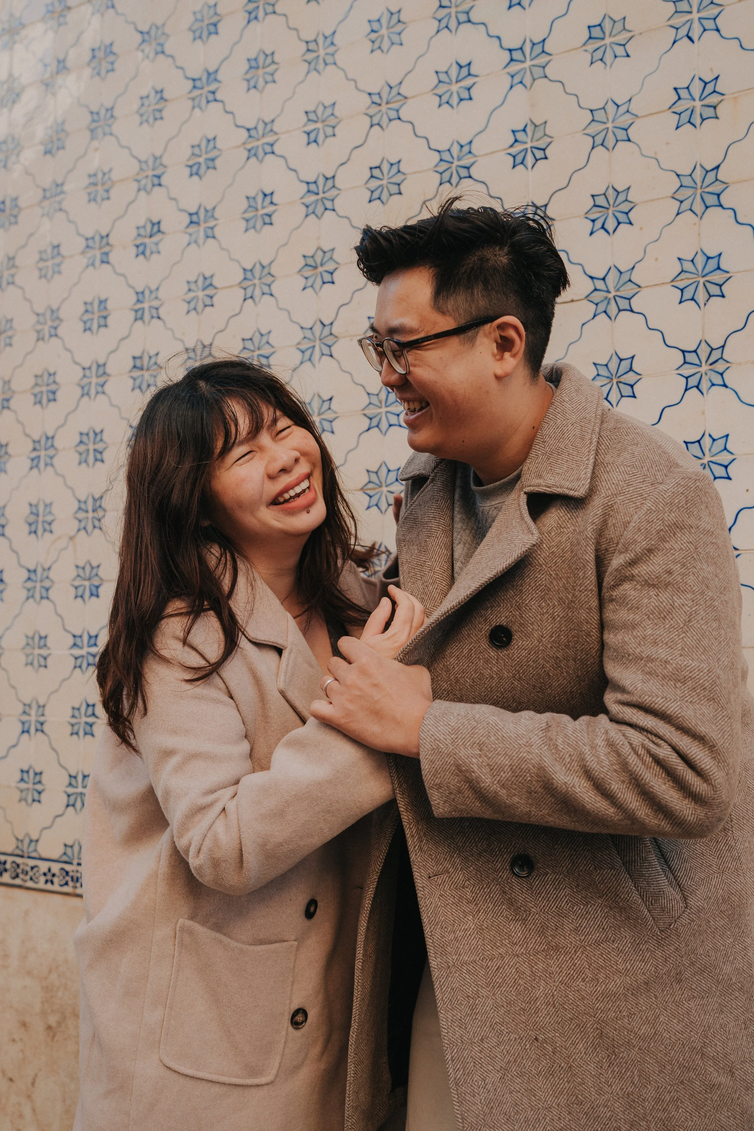 A smiling woman and man share a joyful moment in front of a decorative blue and white tile wall. They are dressed in beige coats, and the woman has long dark hair while the man has short dark hair and glasses.