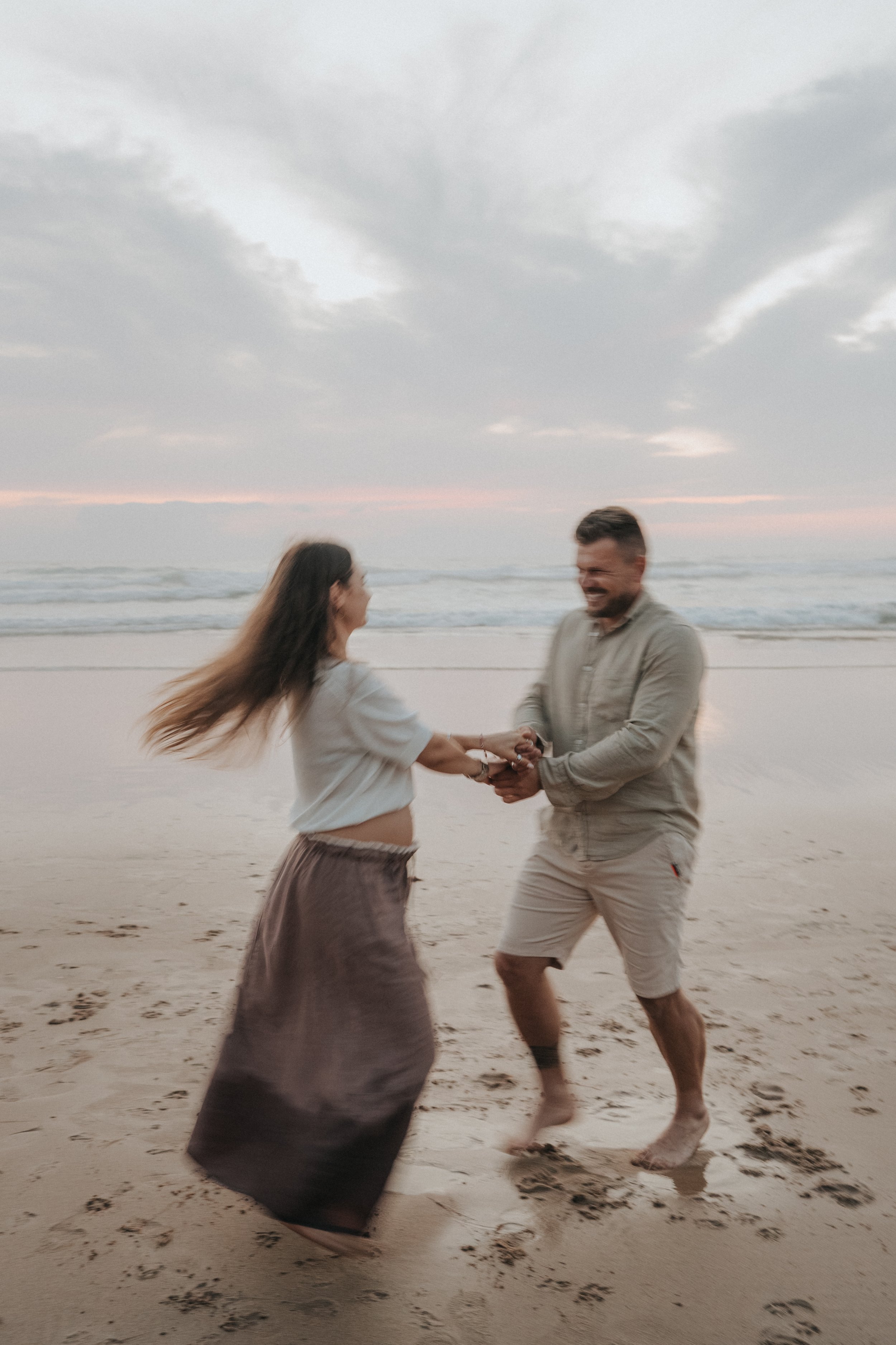 A man and woman dancing or playing on a beach at sunset, holding hands and smiling, with waves and cloudy sky in the background.