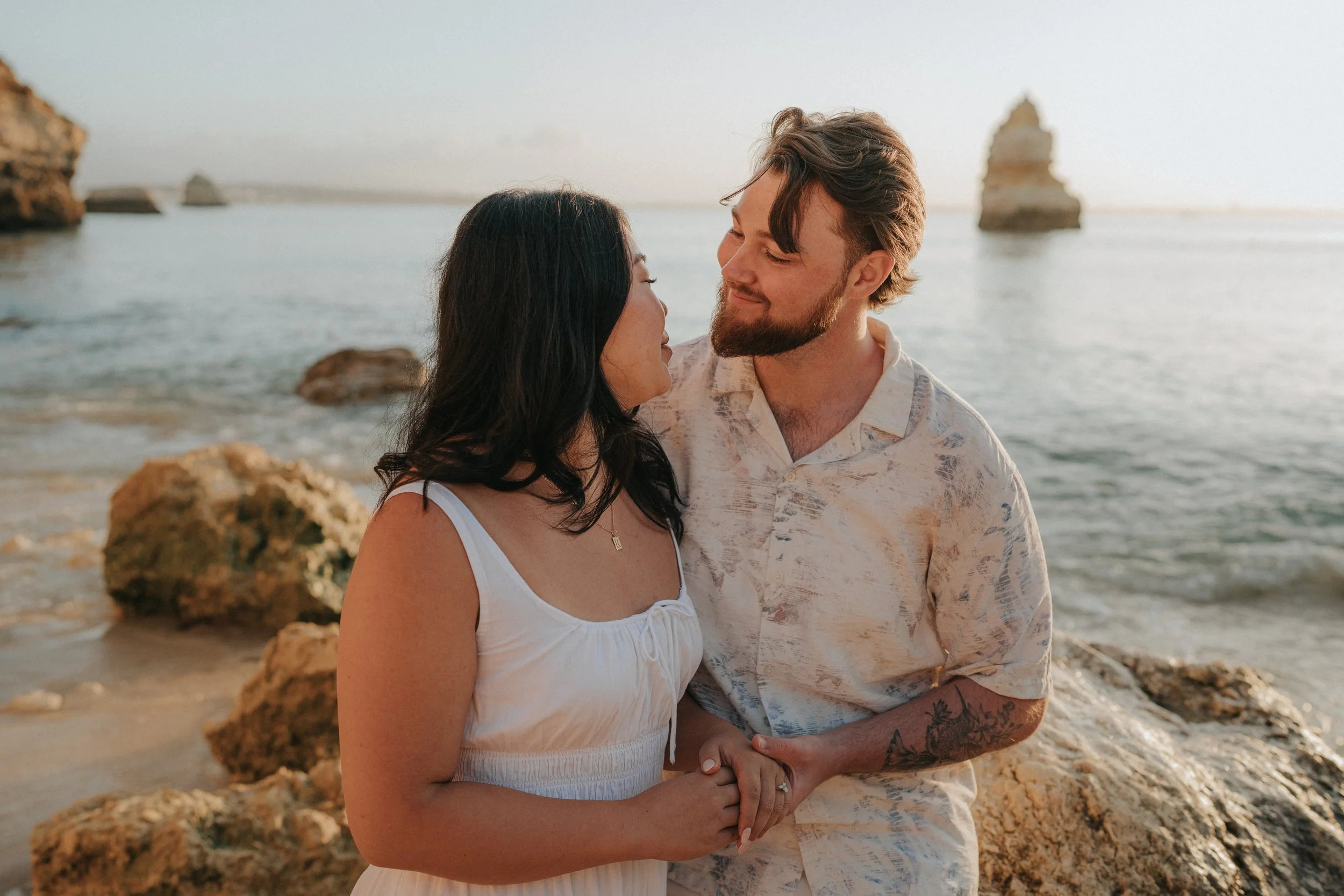 A couple standing close on a rocky beach with the ocean and rock formations in the background during sunset, looking at each other affectionately.