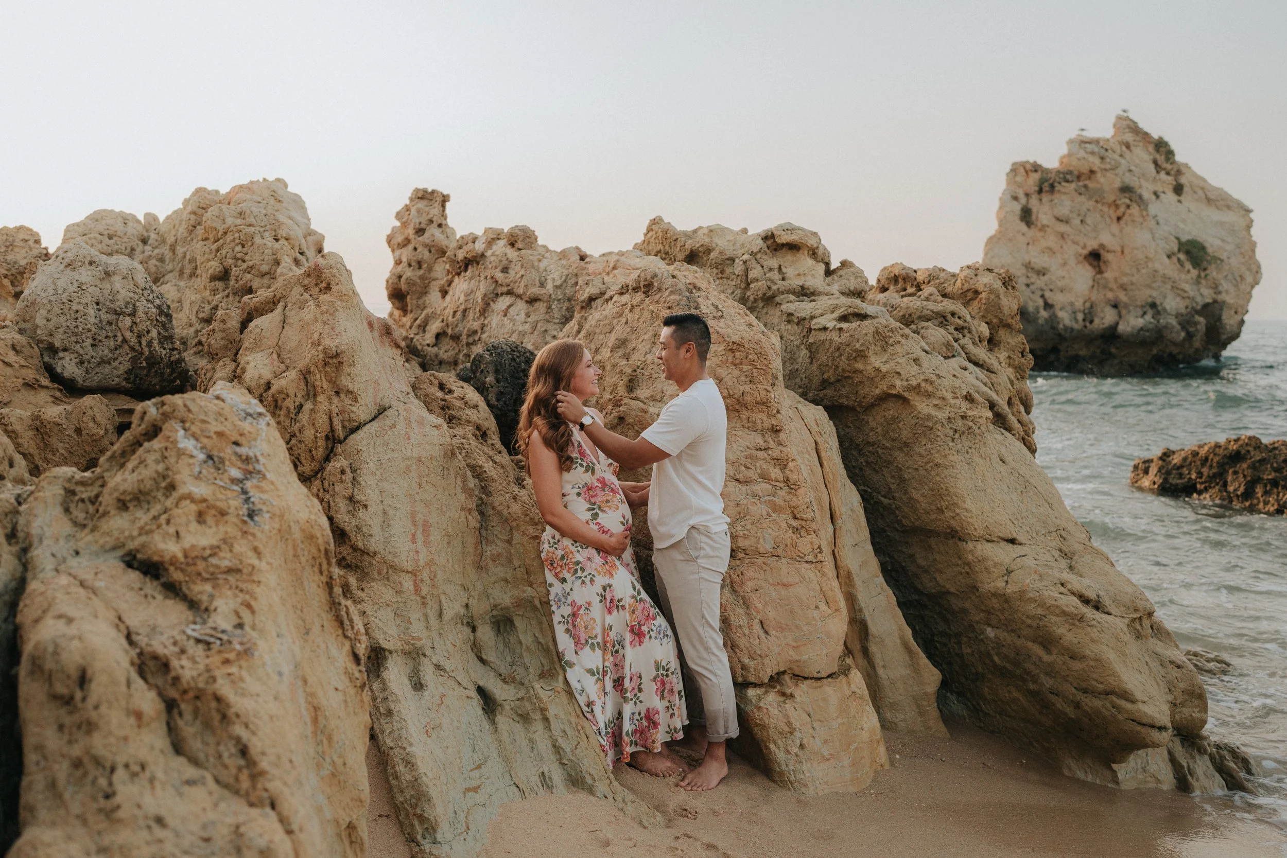 A couple standing on the beach between large rocky formations, with the woman pregnant, gazing at each other near the ocean.