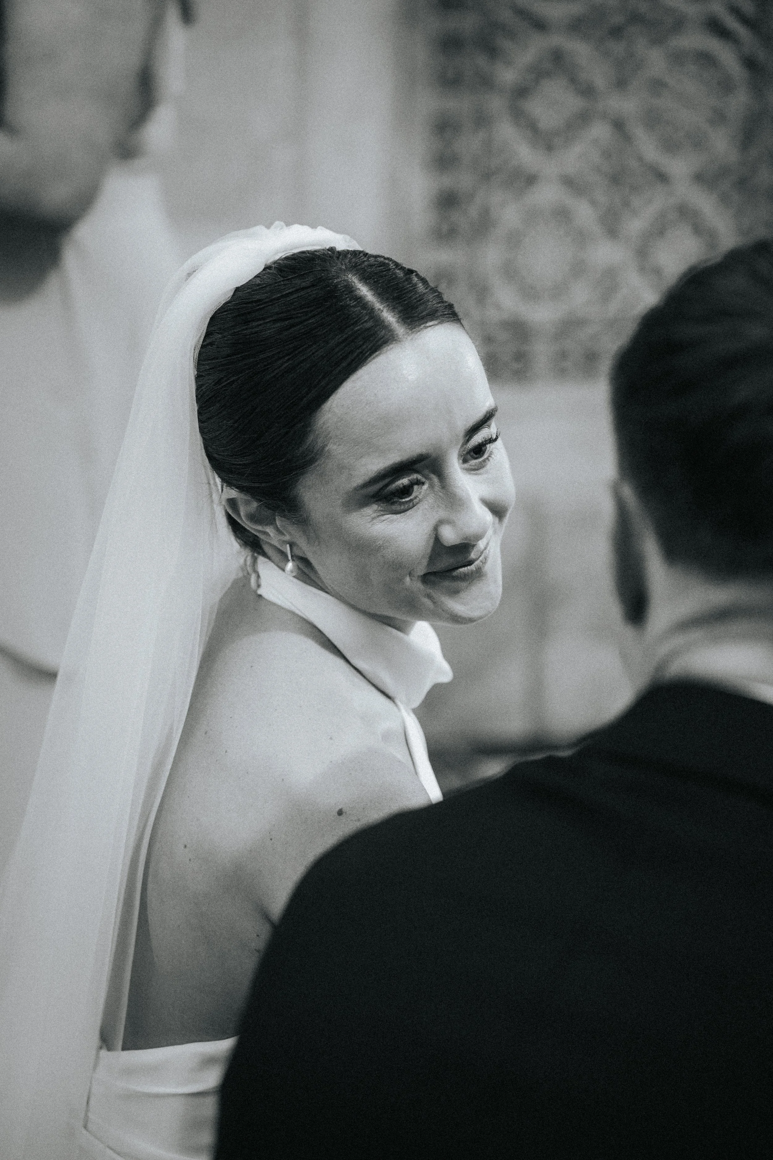 A bride in a white wedding dress with dark hair styled in an elegant updo, wearing a veil, is smiling and looking at someone in a black suit, possibly during a wedding ceremony.