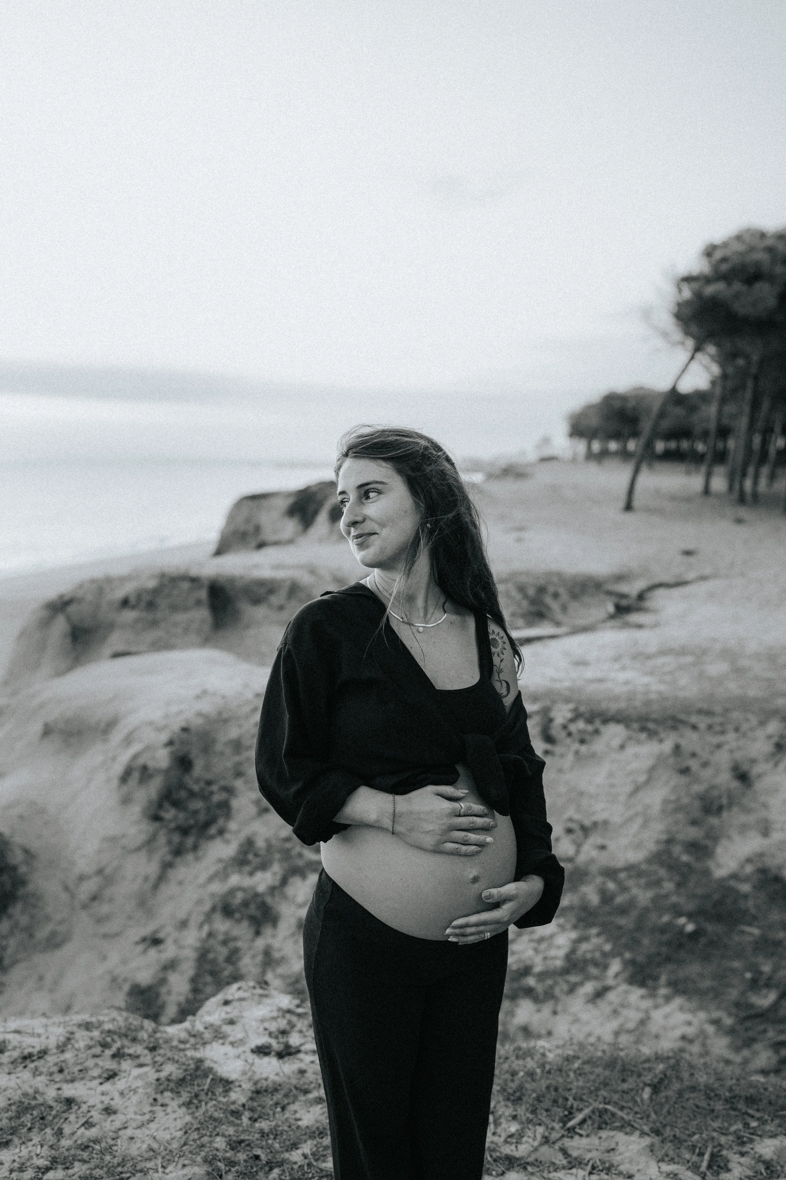 Black and white photo of a pregnant woman standing on a beach, with her hand on her belly and looking off to the side, with dunes and trees in the background.