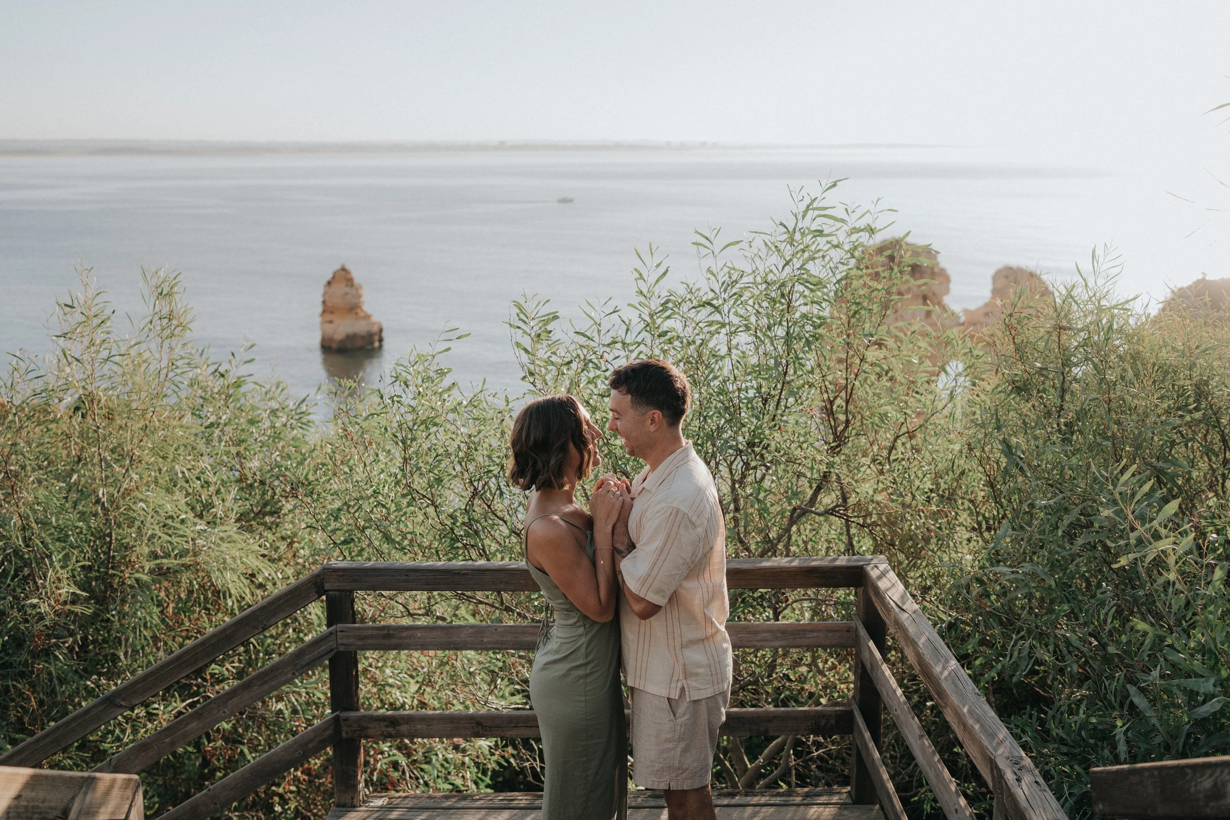 A couple standing close together on a wooden deck near bushes with a body of water and rock formations in the background, engaging in a romantic moment.