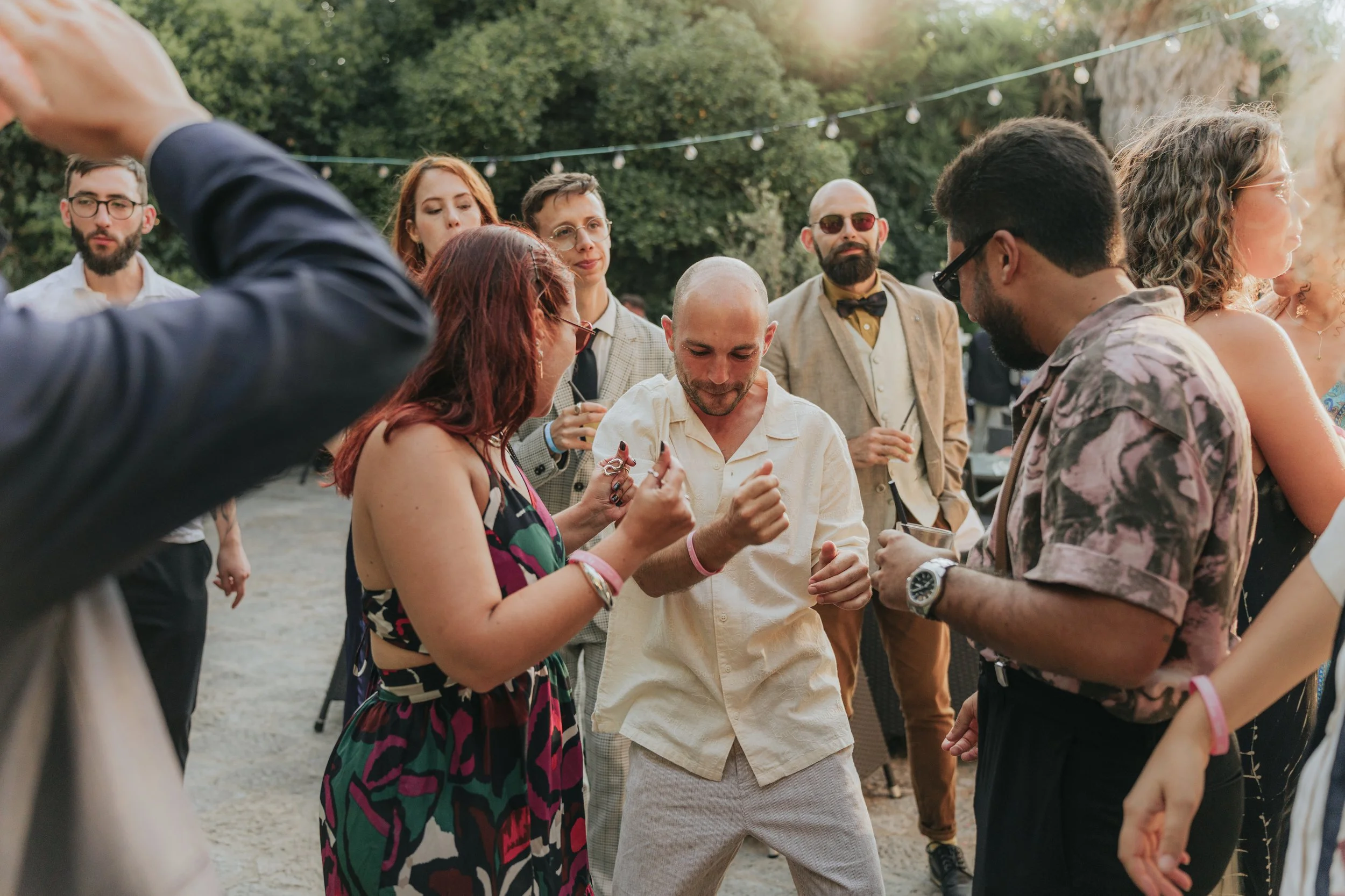Group of people socializing outdoors at a party with string lights overhead, surrounded by trees.