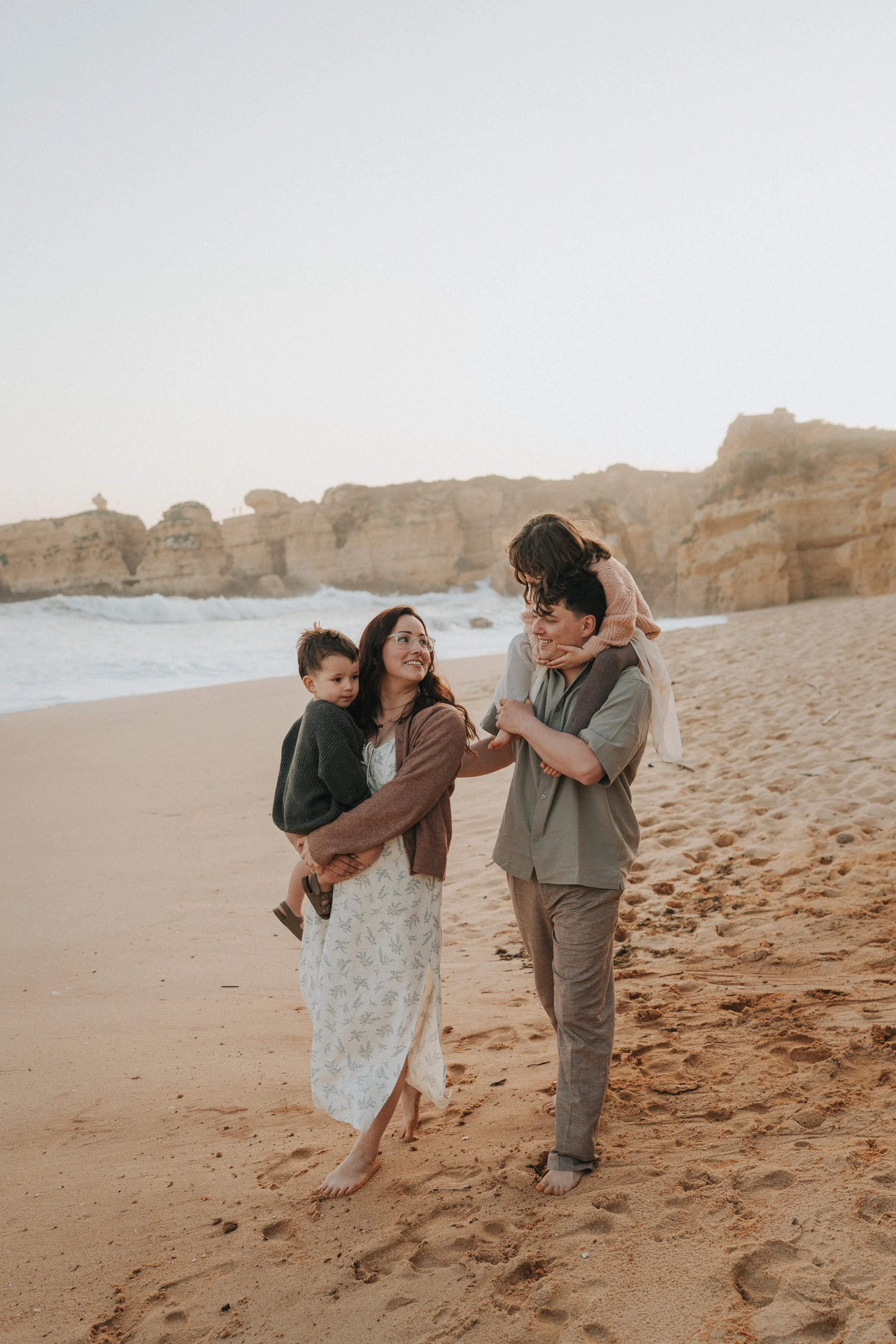 A family of four walking on the beach with cliffs in the background at sunset. The mother is holding a young boy, the father is carrying a girl on his shoulders, and they are all smiling and enjoying each other's company.
