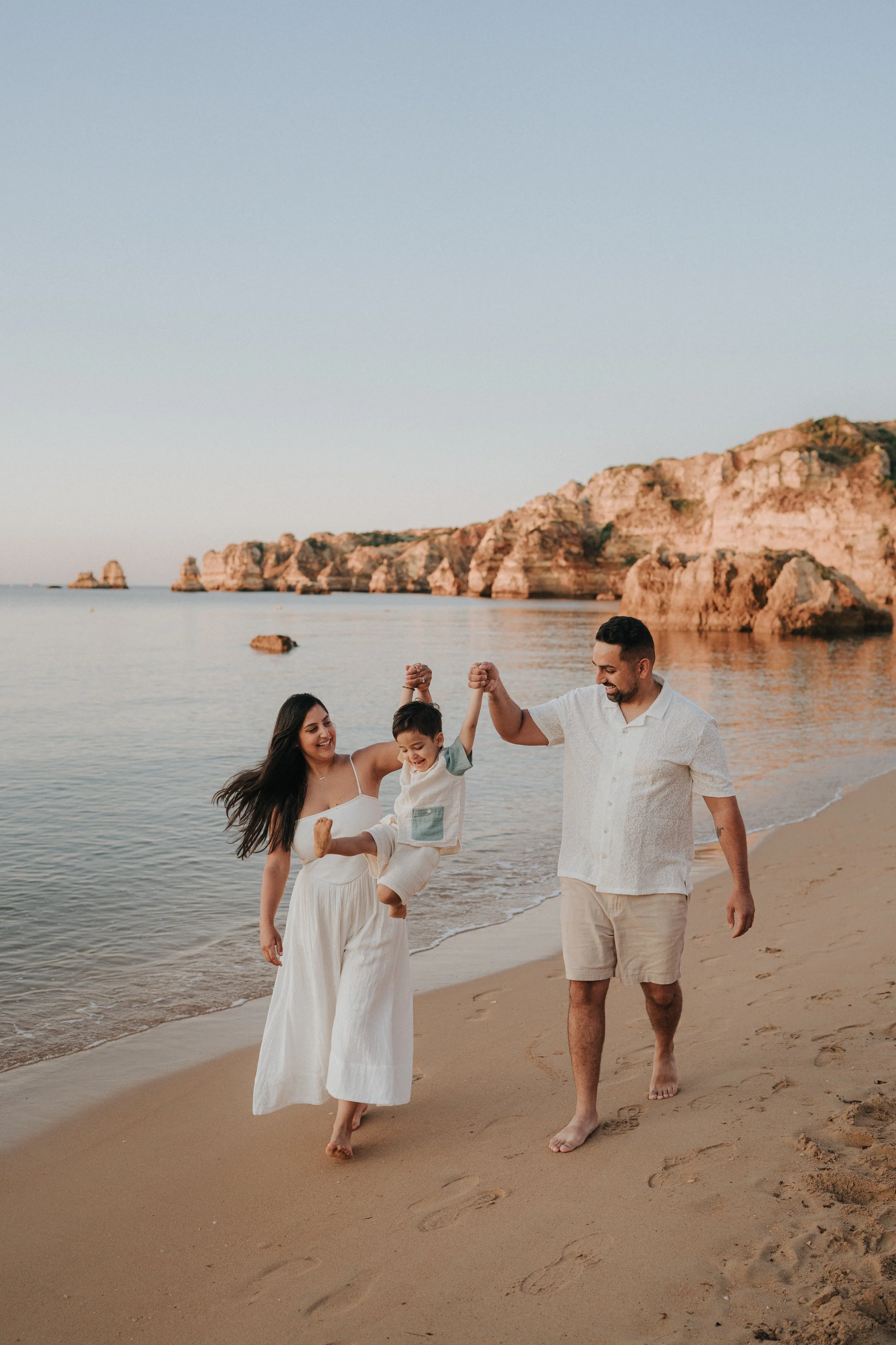 Family of three walking on the beach, with the father lifting the young boy, all smiling, with rocky cliffs in the background during sunset.