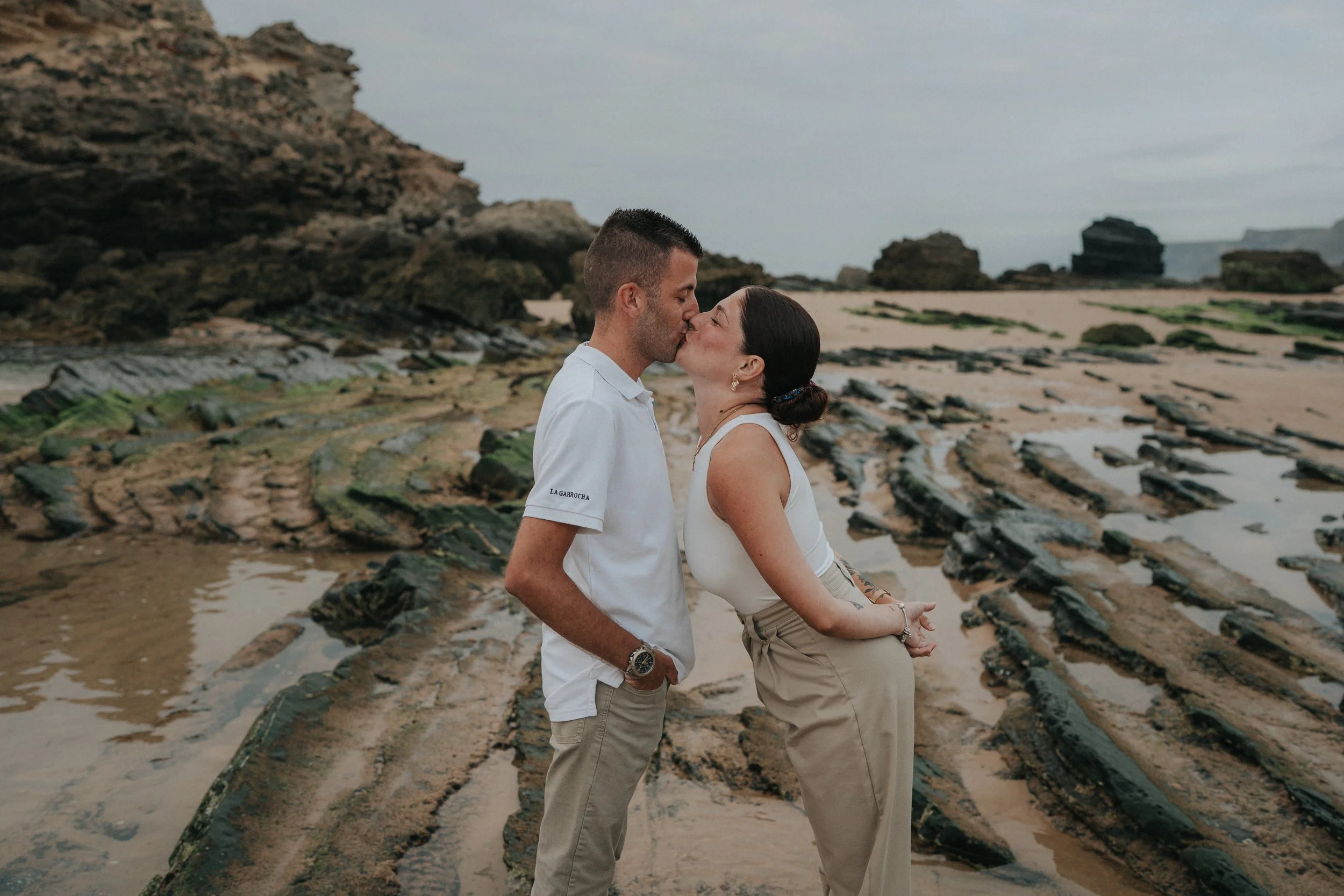 A couple sharing a kiss on a rocky beach with cliffs and tide pools in the background.