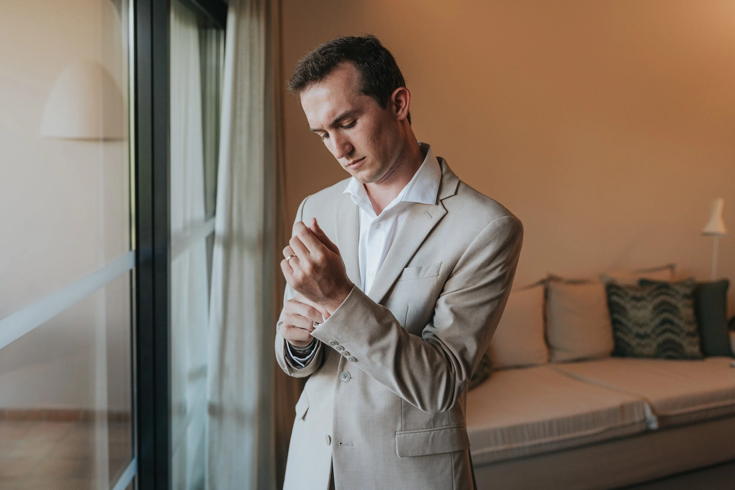 A man in a beige suit adjusting his shirt cuff in a room with a bed and window with curtains, soft lighting, and a lamp on the wall.