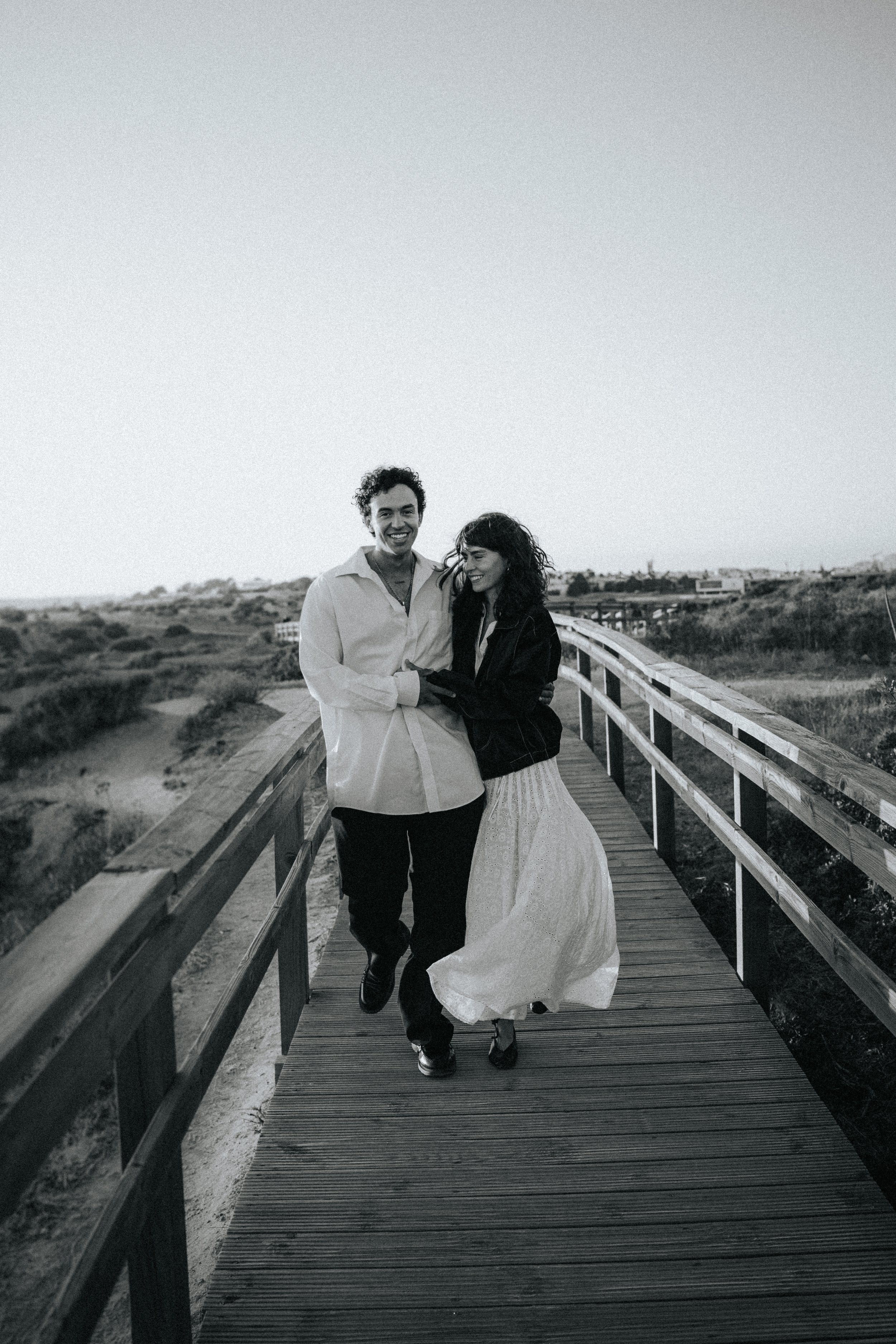 A black-and-white photo of a happy young man and woman walking hand in hand on a wooden bridge outdoors, with scenic natural landscape and distant buildings in the background.