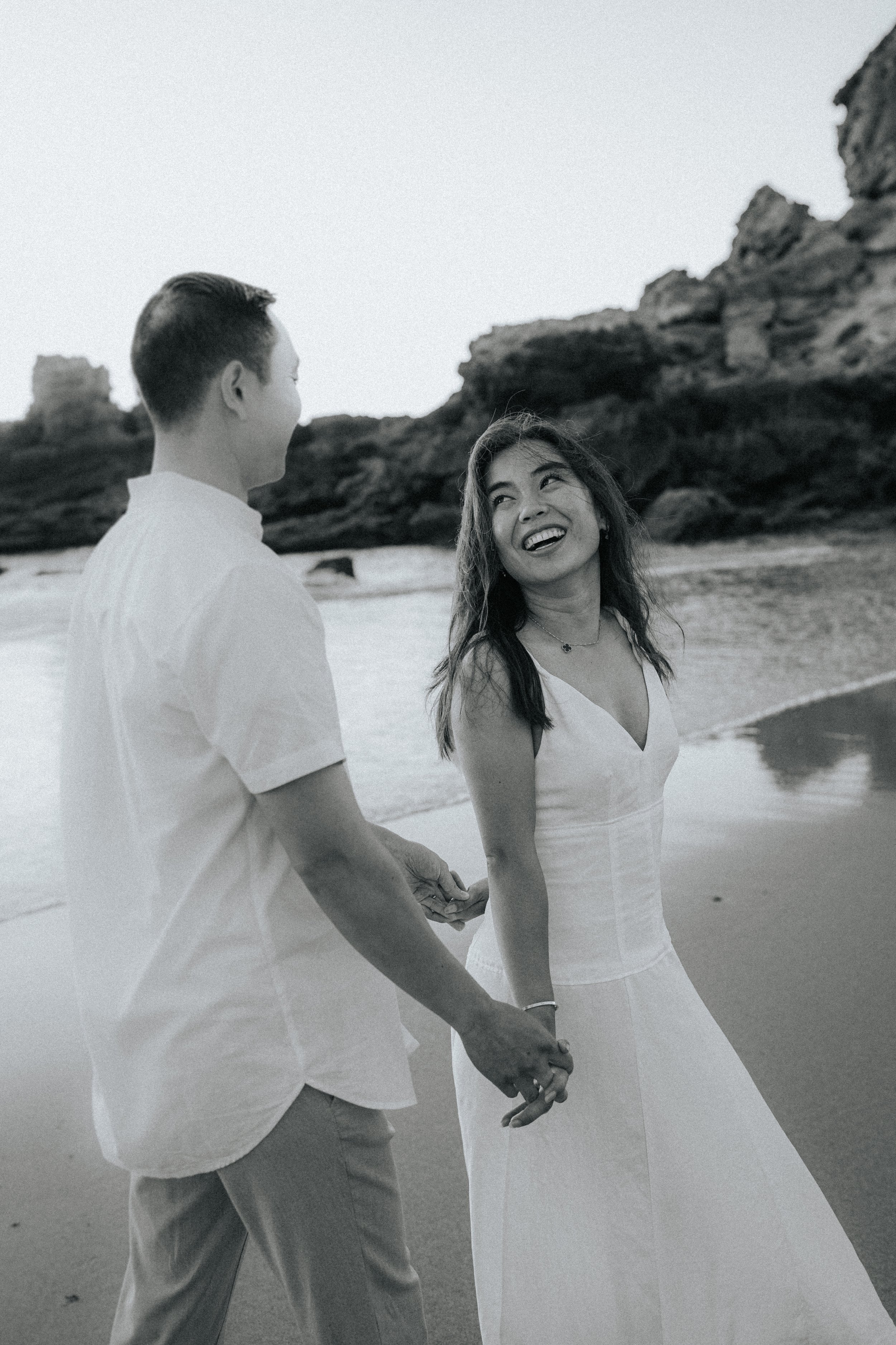 A couple holding hands on the beach, dressed in white, with the woman smiling and looking at the man. Rock formations are visible in the background.